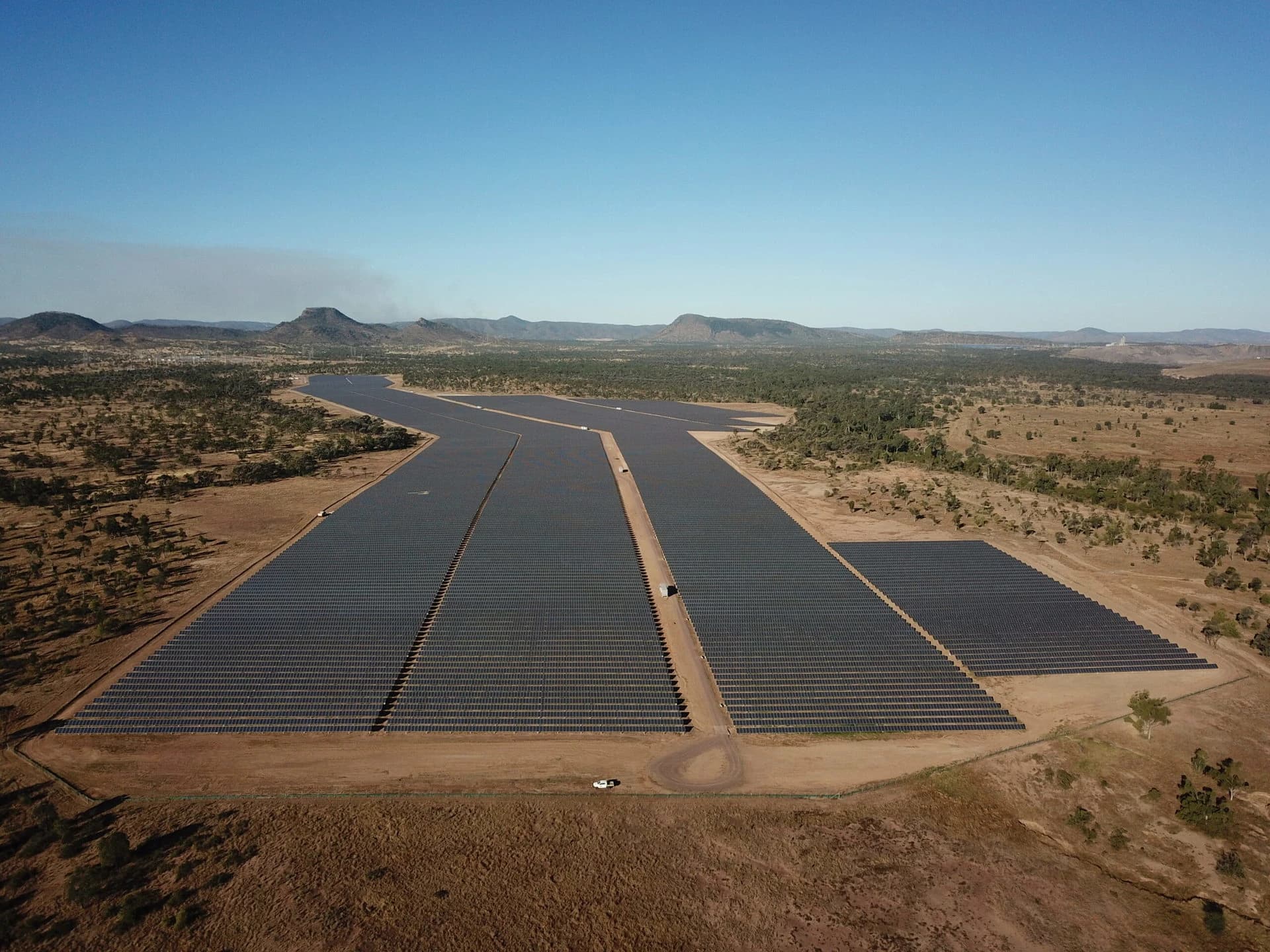 Vue aérienne d'un vaste parc solaire composé de milliers de panneaux bleus alignés sur un paysage désertique ocre. Un canal bleu, une route et quelques bâtiments bordent le site sous un ciel clair et ensoleillé.