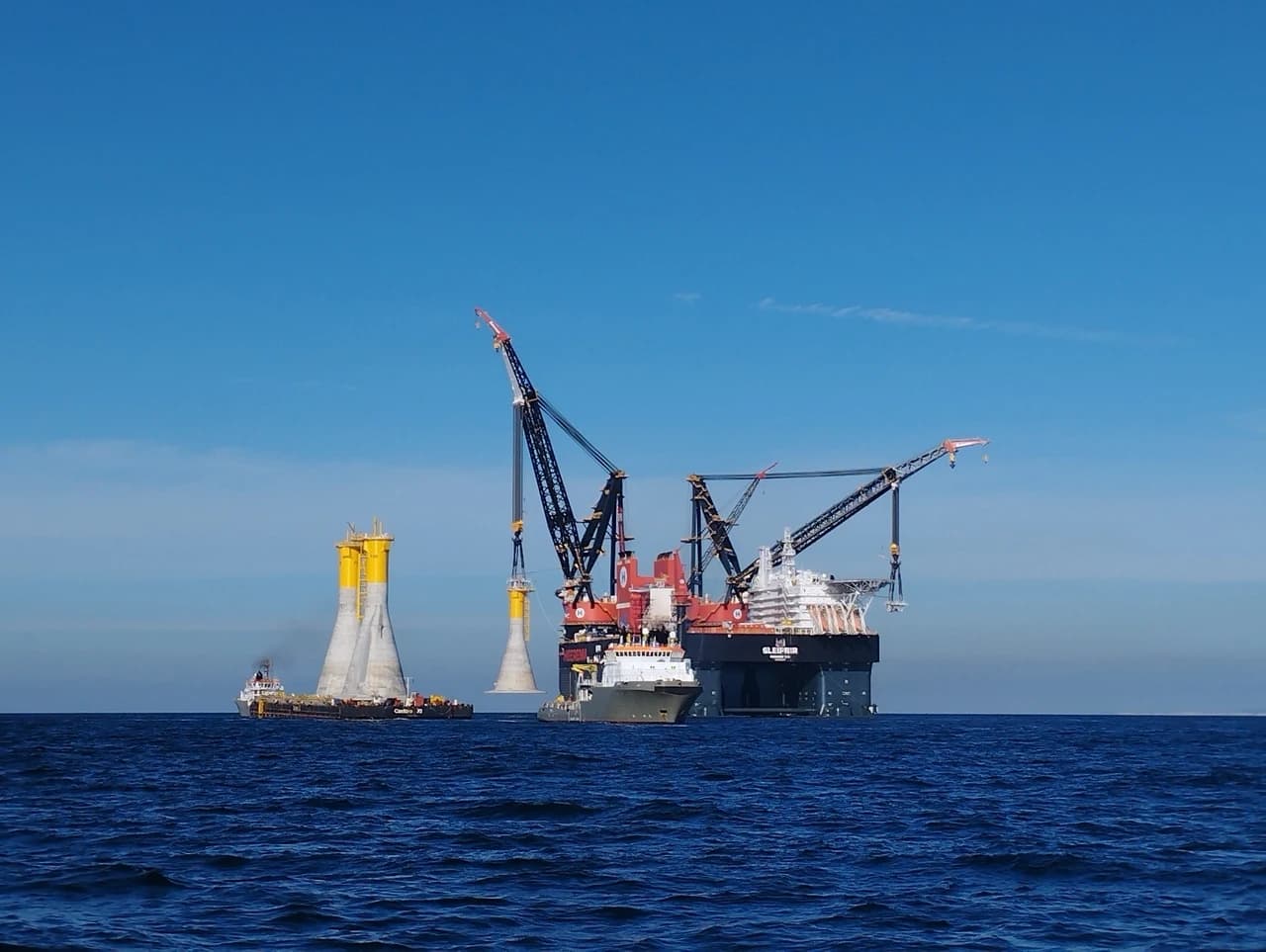 Grand navire-grue Sleipnir, rouge et noir, soulevant un composant de fondation d'éolienne conique gris et jaune en mer, à côté d'une barge transportant deux structures similaires sous un ciel bleu clair.