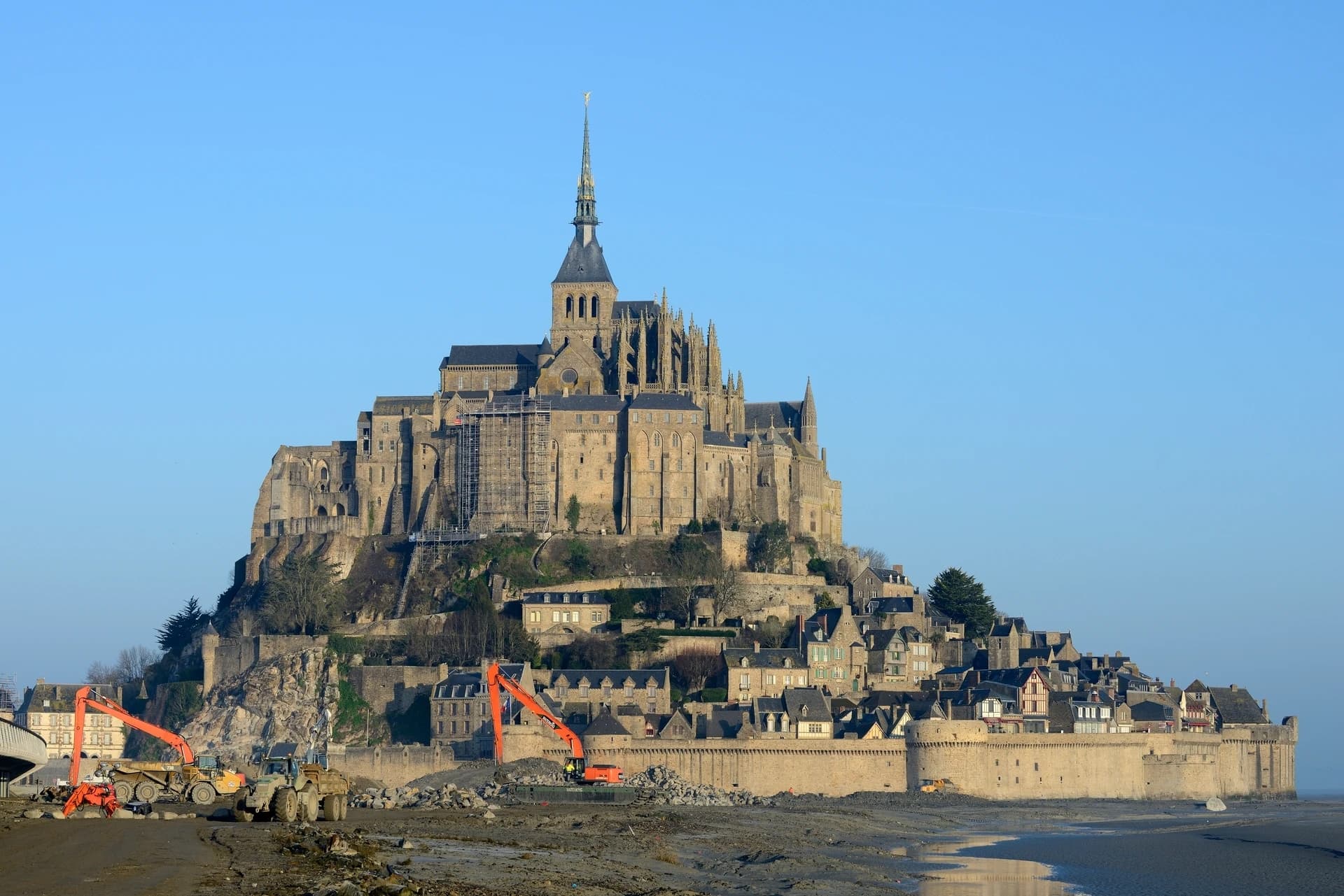 Le Mont Saint-Michel, avec son imposante abbaye et son village médiéval en pierre, se dresse sous un ciel bleu clair. Au premier plan, des engins de chantier orange travaillent sur les étendues de sable et de boue à marée basse, tandis que des échafaudages sont visibles sur une partie de l'abbaye.