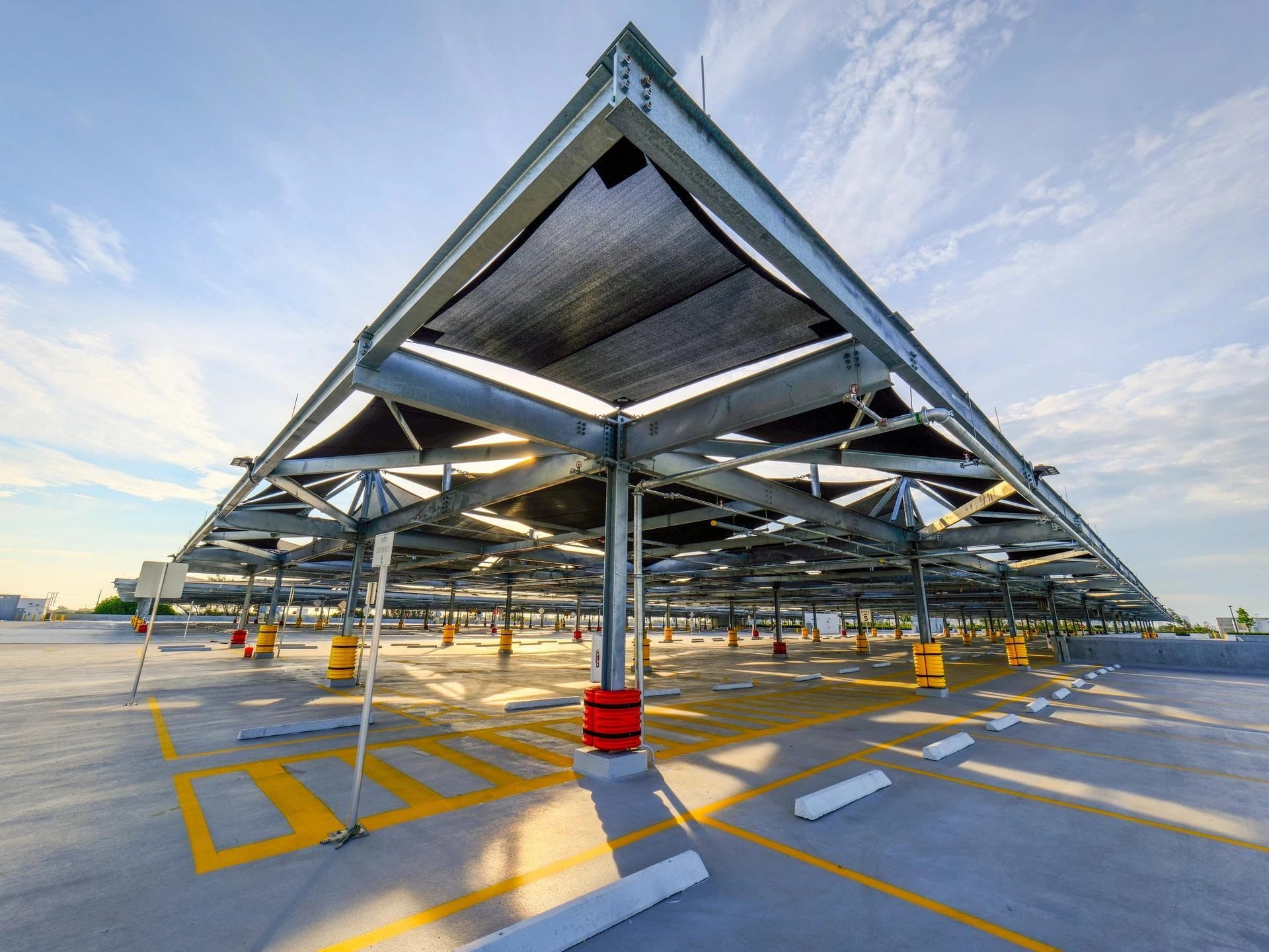 Grand parking extérieur vide avec une structure métallique grise et des panneaux d'ombrage noirs triangulaires sous un ciel bleu clair. Le sol en béton gris est marqué de lignes de stationnement jaunes vives et de butées blanches, baigné d'une lumière dorée créant de longues ombres.