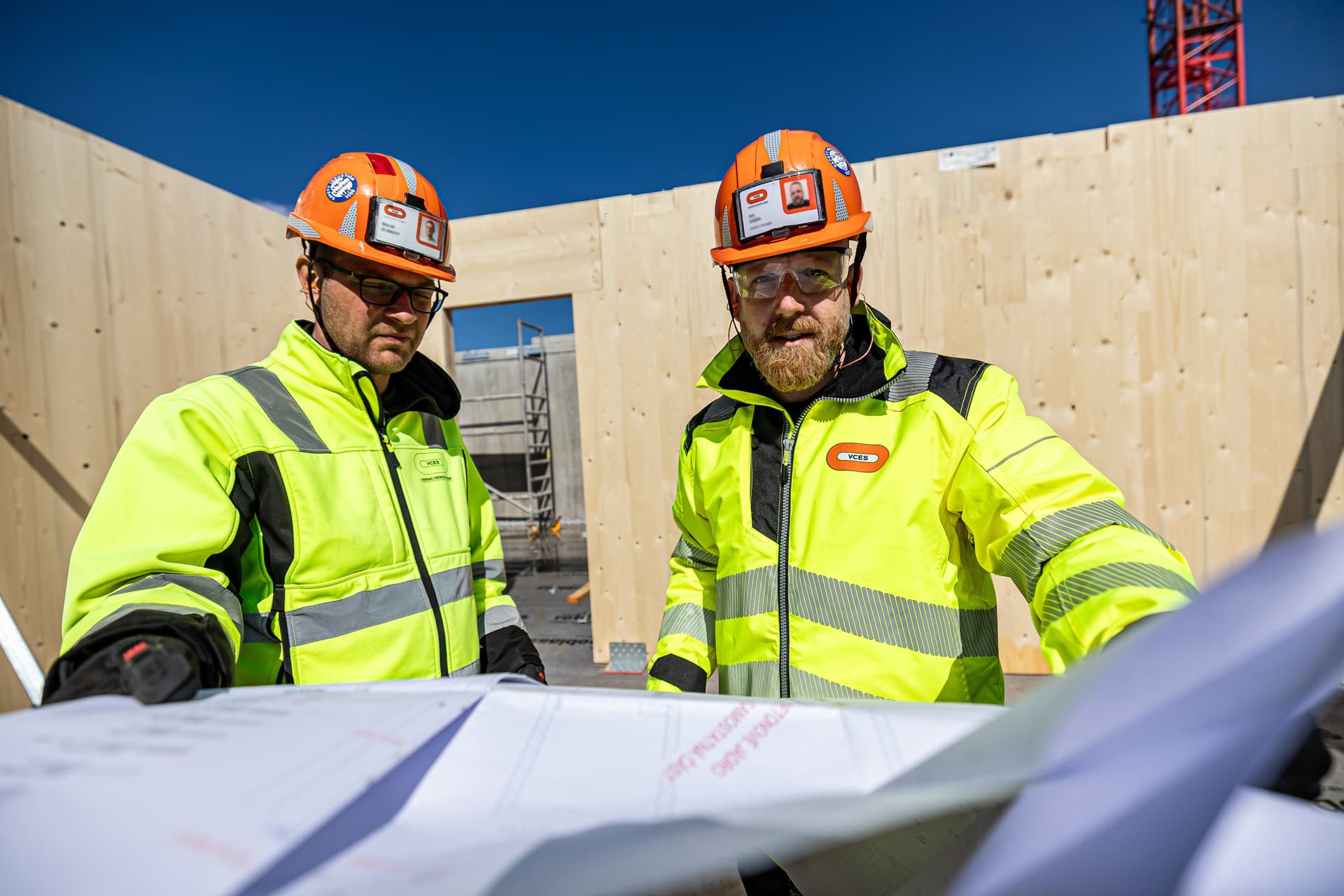 Deux hommes en vestes de sécurité jaune fluo et casques orange examinent des plans sur un chantier de construction en bois, sous un ciel bleu clair.