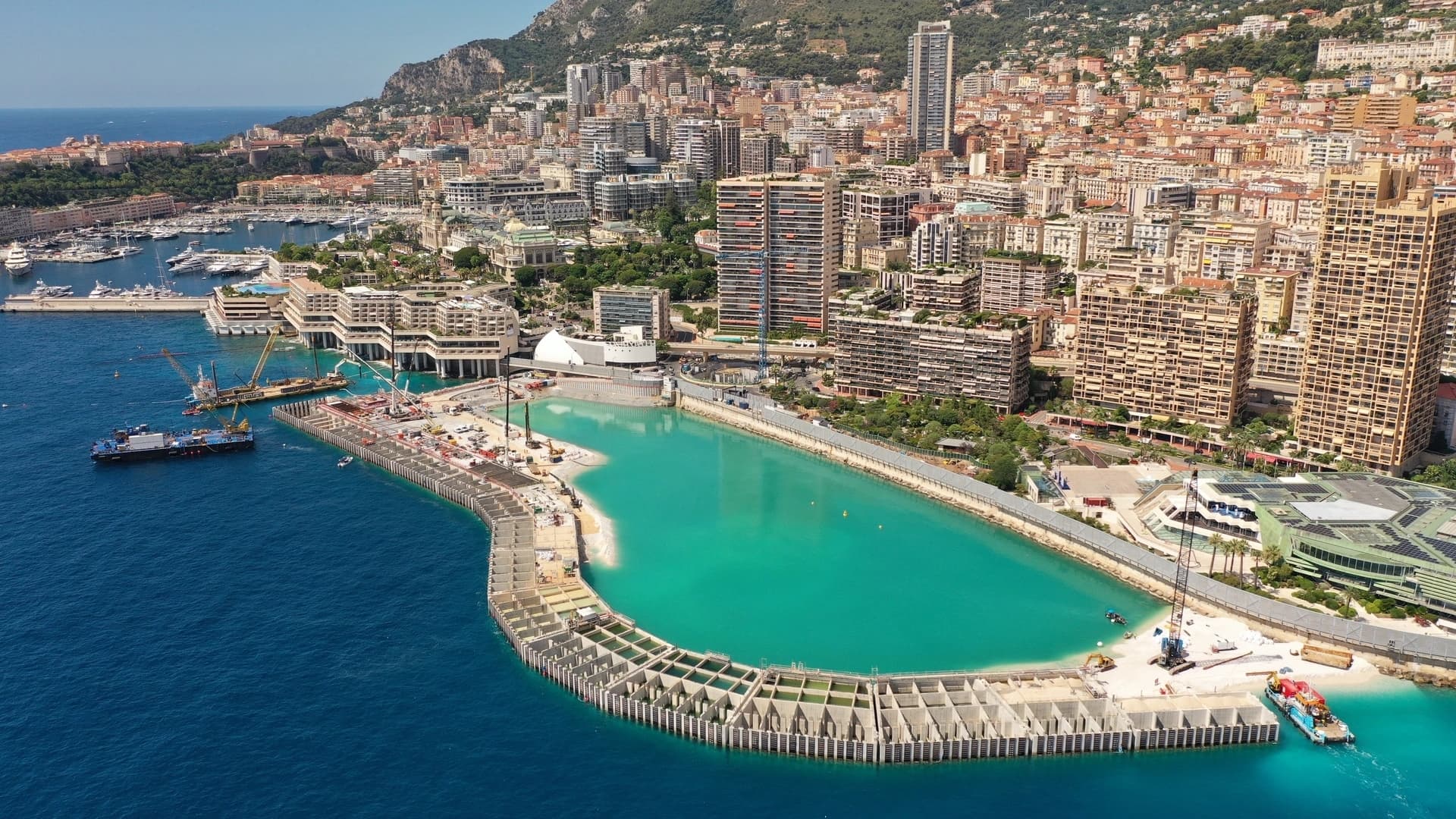 Vue aérienne de Monaco avec son dense paysage urbain sur des collines, un port rempli de yachts, et un grand projet d'extension côtière en cours, caractérisé par une étendue d'eau turquoise et des structures de béton incurvées.