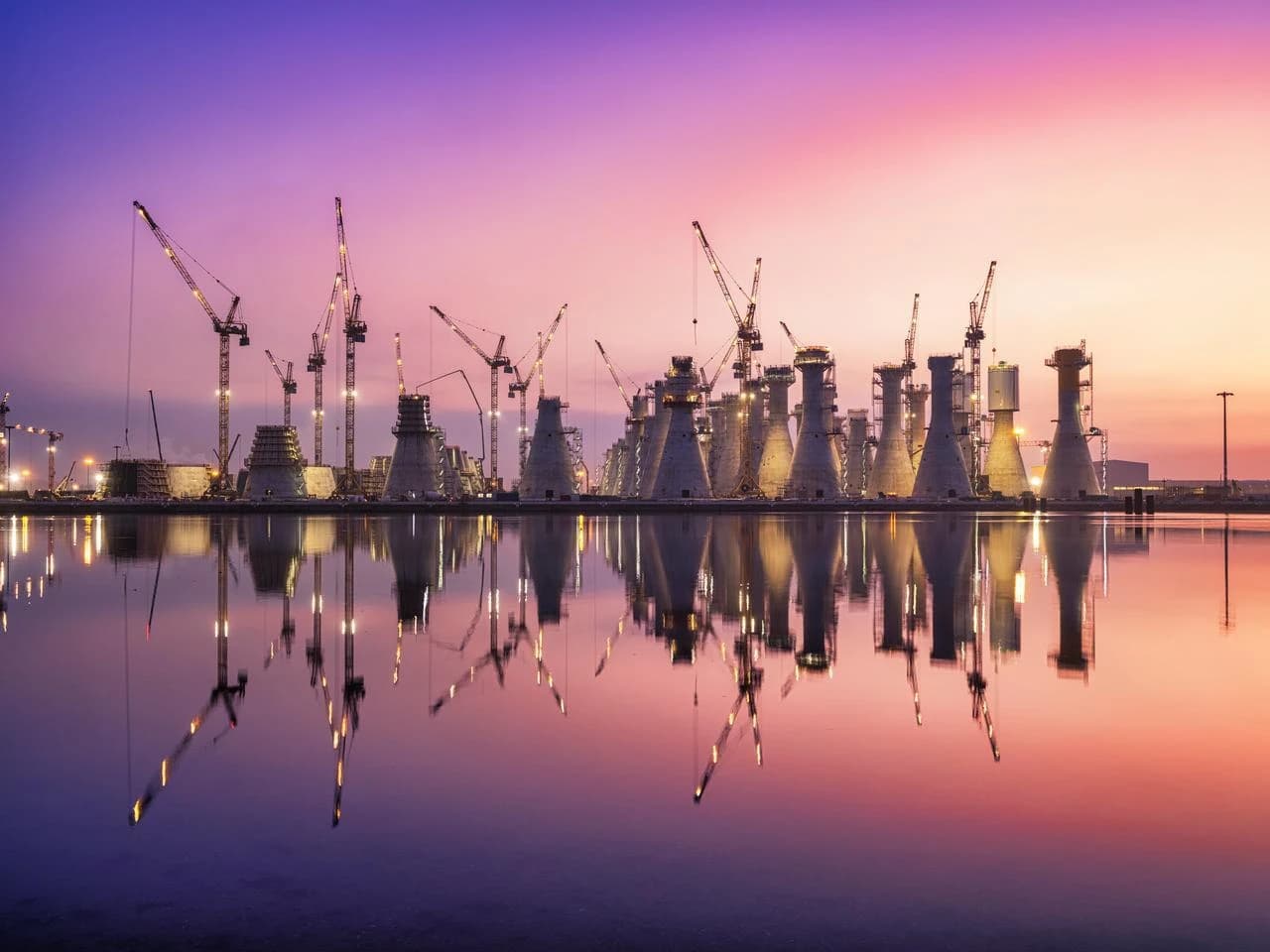Un site de construction industriel avec de nombreuses structures coniques et grues en cours d'édification, parfaitement reflété dans l'eau sous un ciel de crépuscule aux dégradés de violet, rose et orange.