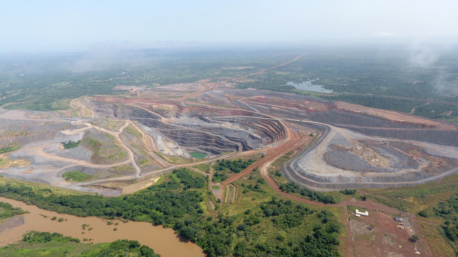 Vue aérienne d'une vaste mine à ciel ouvert présentant des terrasses concentriques de roche grise et terre rougeâtre, contrastant avec les forêts tropicales verdoyantes et les eaux boueuses en premier plan. Des routes d'accès serpentent à travers le site industriel sous un ciel clair mais légèrement brumeux.
