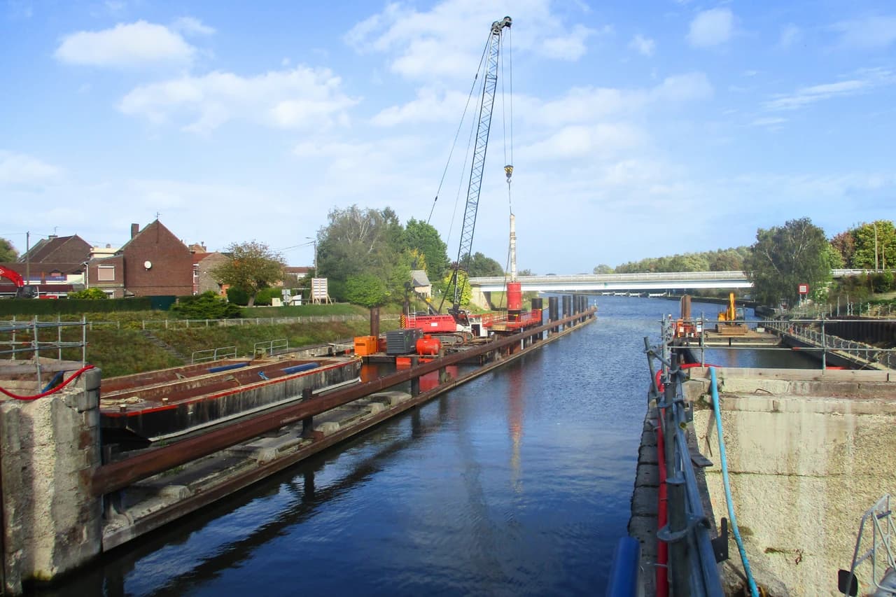 Une grue rouge imposante sur une barge opère sur un canal bleu, avec des maisons en briques sur la rive gauche et un pont blanc à l'horizon, sous un ciel clair nuageux.