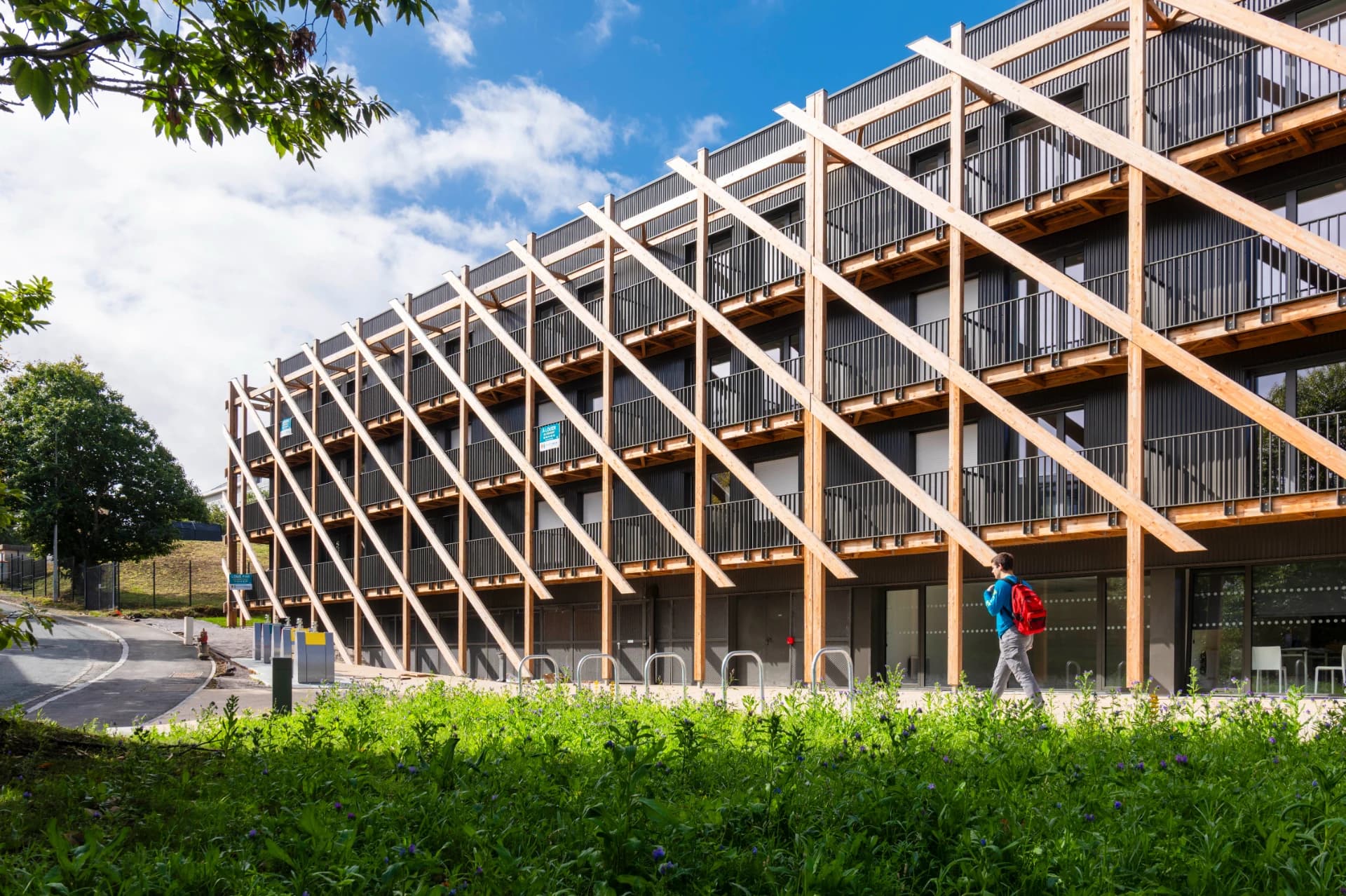 Bâtiment moderne à la façade sombre avec un treillis diagonal en bois clair et des balcons, sous un ciel bleu nuageux. Au premier plan, une pelouse verte où une personne marche le long de l'édifice.
