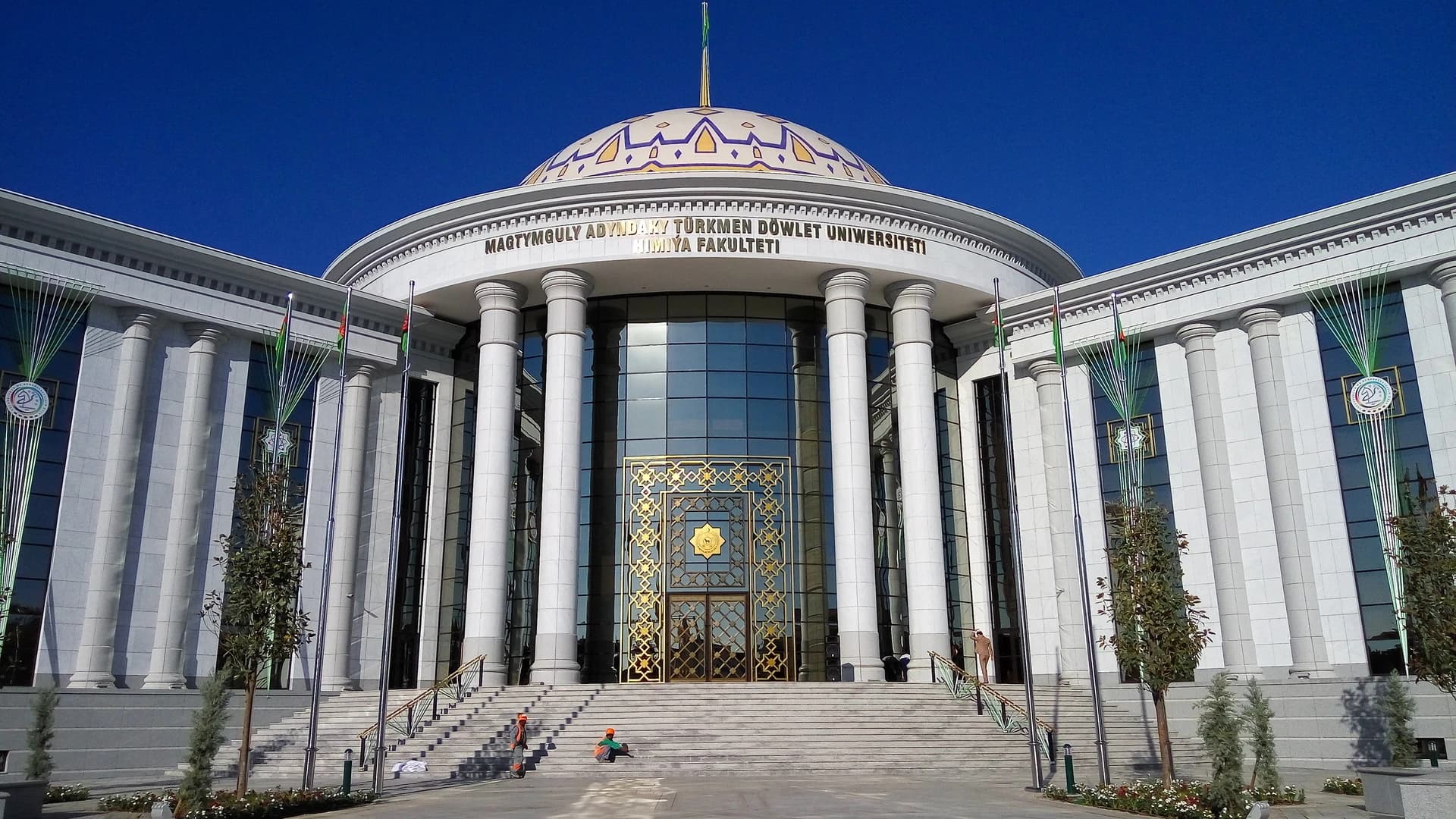 Façade majestueuse de la Faculté de Chimie de l'Université d'État turkmène Magtymguly, un grand bâtiment blanc de style néoclassique avec de hautes colonnes et un dôme doré et bleu. L'entrée principale est un mur de verre orné d'un cadre doré complexe, sous un ciel bleu clair.