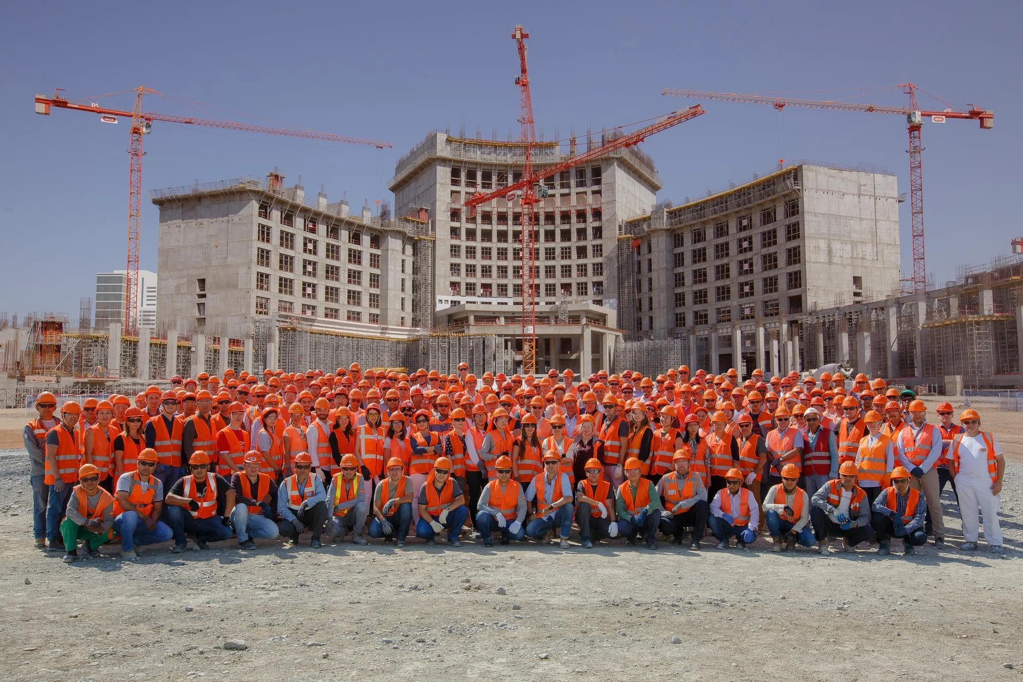 Un grand groupe d'ouvriers et de personnel portant des gilets et casques de sécurité orange posent sur un chantier, avec de grands bâtiments en construction et plusieurs grues rouges en arrière-plan sous un ciel bleu clair.