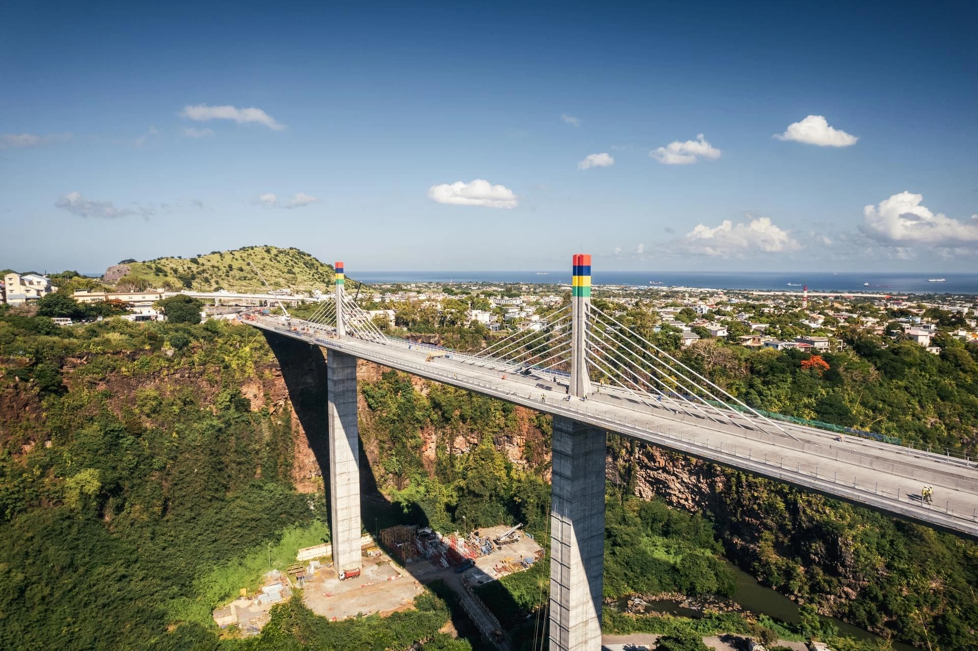 Vue aérienne d'un pont à haubans moderne avec des pylônes gris surmontés de drapeaux mauriciens colorés, traversant une profonde vallée verdoyante. En arrière-plan, une ville s'étend vers l'océan bleu sous un ciel clair.
