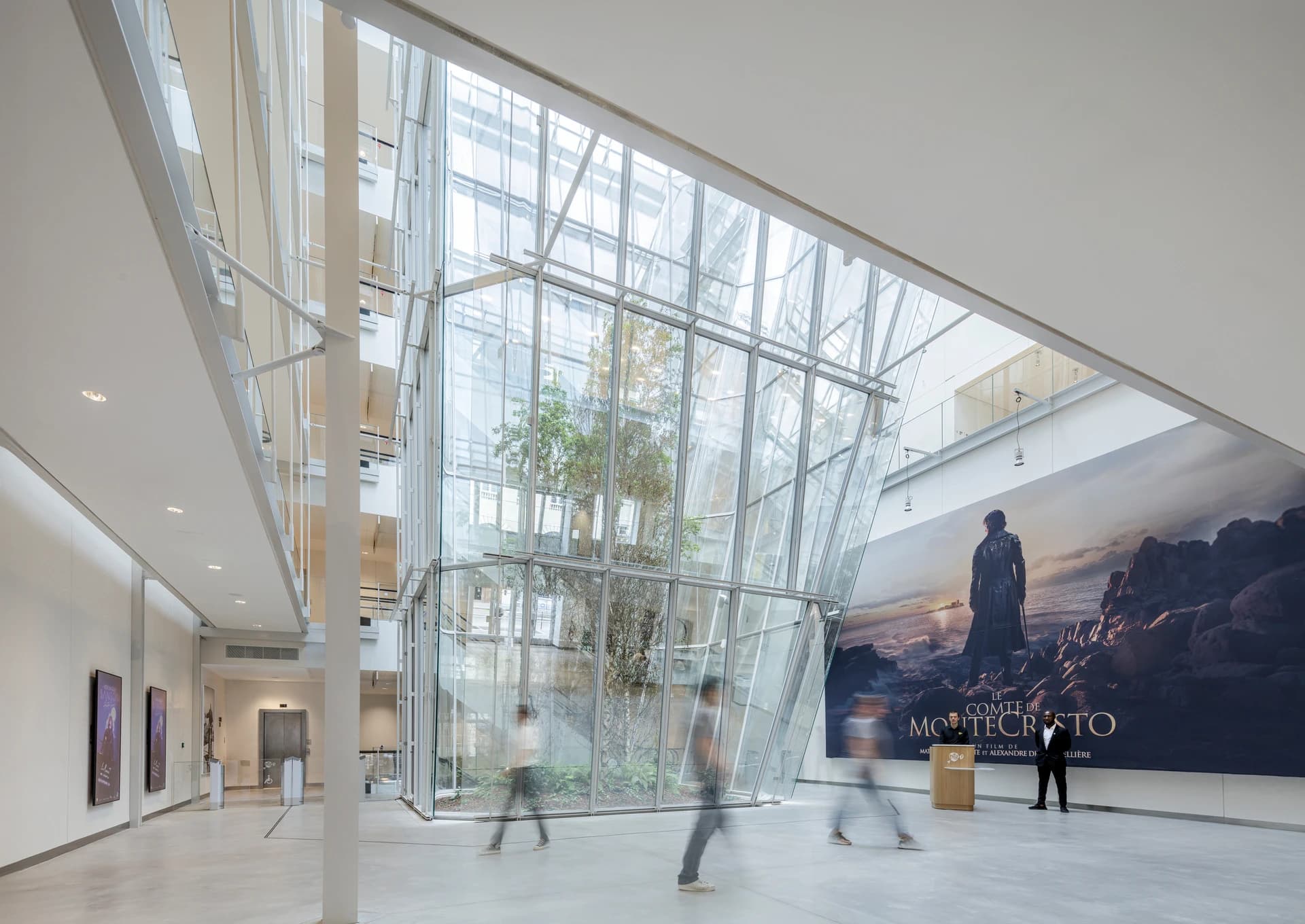 Un vaste hall moderne et lumineux est dominé par une grande verrière centrale abritant un arbre et un immense poster mural du film Le Comte de Monte-Cristo. Des personnes aux mouvements flous traversent cet espace contemporain aux murs blancs et au sol gris.