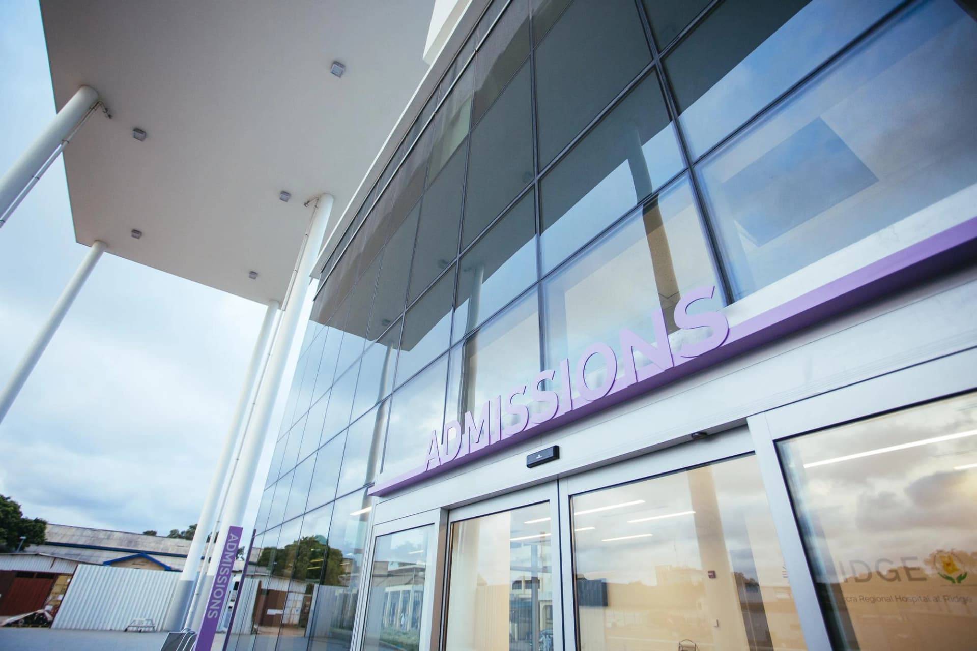 Entrée d'un hôpital moderne en verre avec un panneau 'ADMISSIONS' violet et des colonnes de marquise blanches.