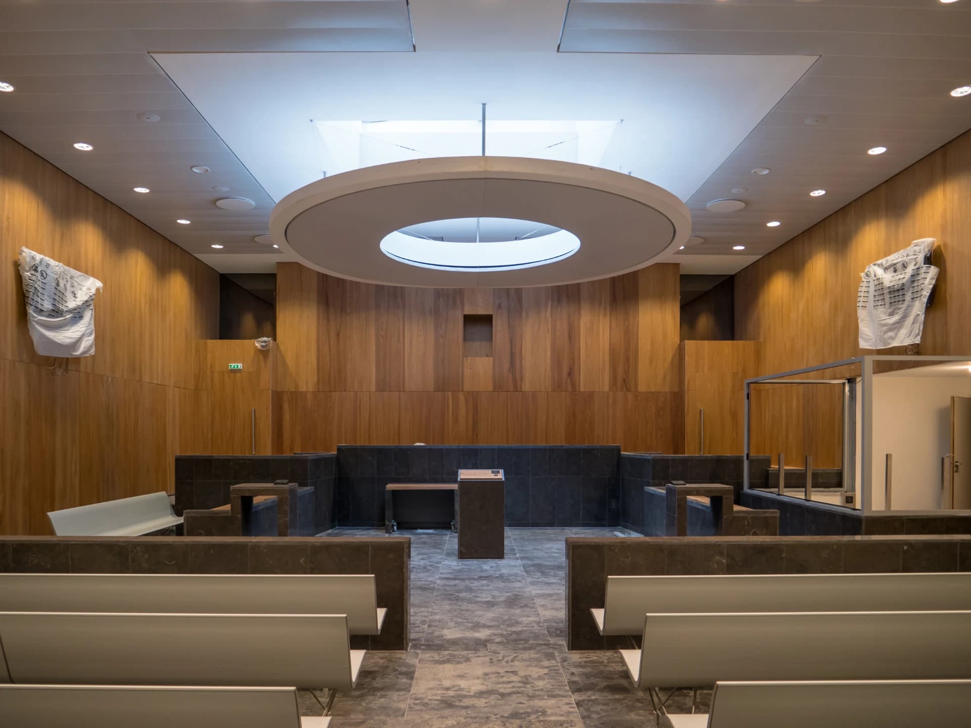 Intérieur d'une salle d'audience moderne et vide, avec des murs en lambris de bois clair et des bancs gris clair pour le public. L'estrade et le pupitre sont en pierre sombre sous un grand luminaire circulaire au plafond blanc lumineux.