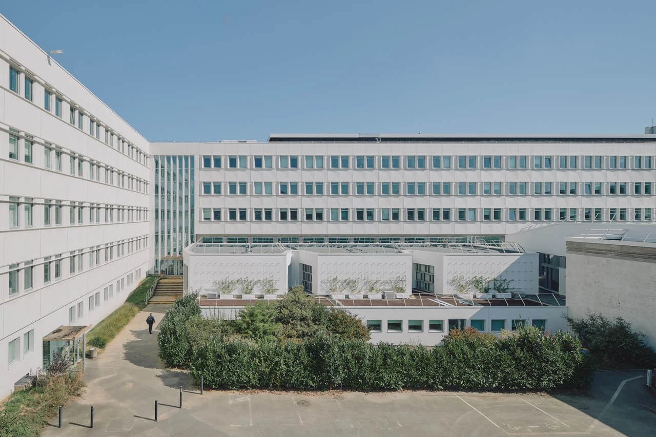 Vue extérieure d'un grand bâtiment blanc moderne avec de nombreuses fenêtres, une cour verdoyante et un ciel bleu clair, avec une personne marchant.