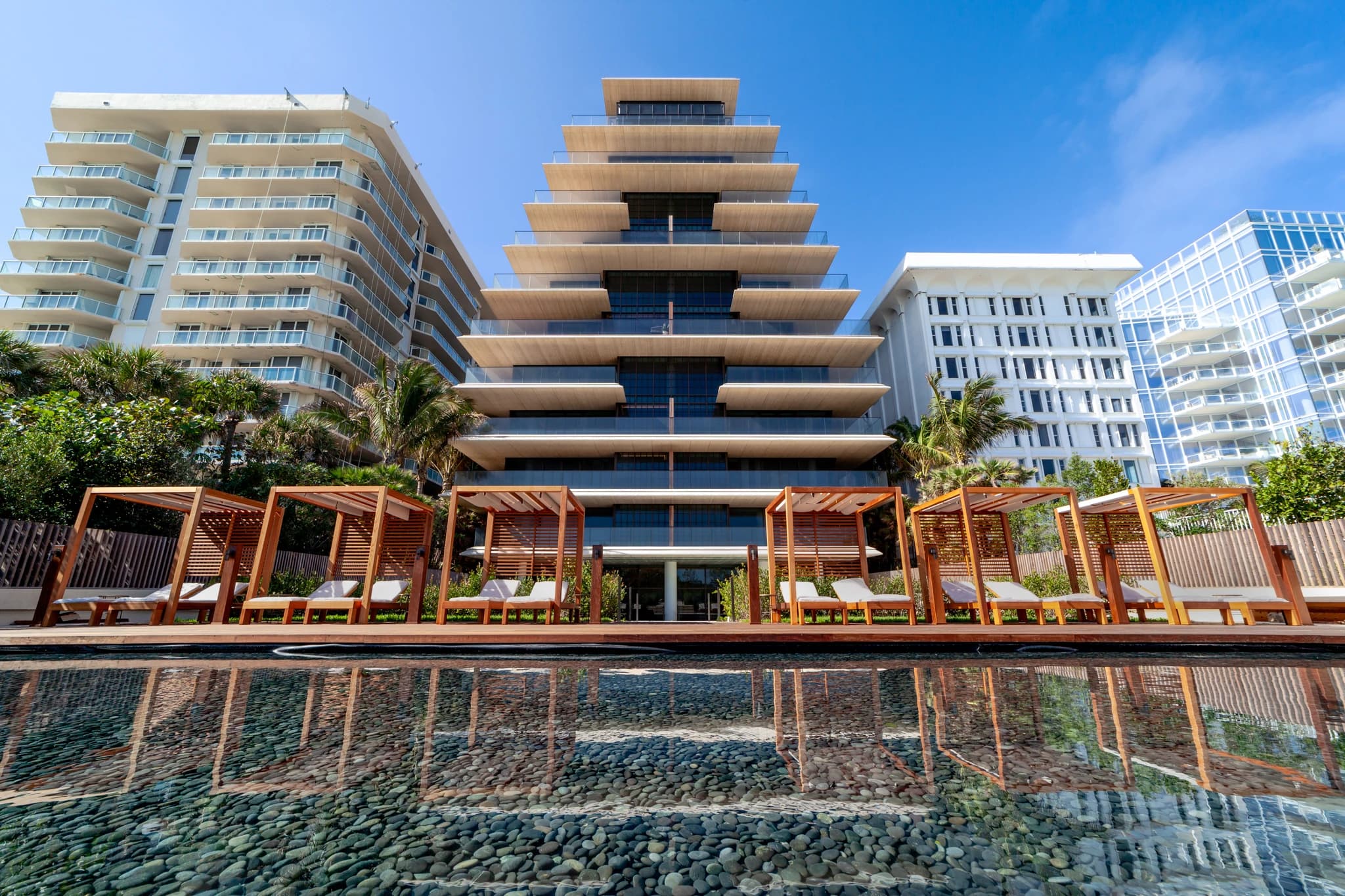 Un immeuble moderne central en terrasses de pierre claire et de verre se dresse sous un ciel bleu, entouré d'autres gratte-ciel. Au premier plan, une piscine à fond de galets reflète l'architecture et est bordée de cabanas en bois avec des chaises longues blanches.