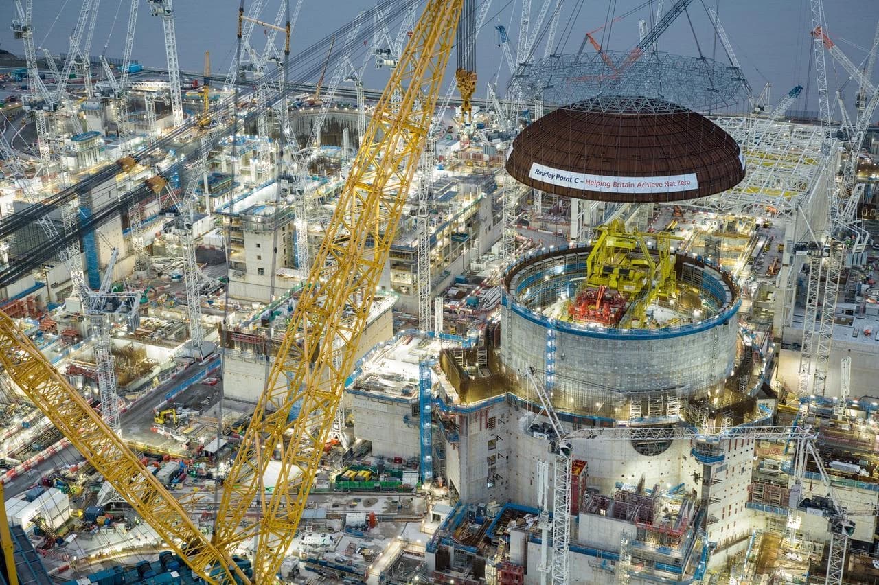 Vue aérienne d'un immense chantier de construction nocturne, dominé par de nombreuses grues blanches et une grue jaune géante soulevant une coupole brune marquée "Hinkley Point C - Helping Britain Achieve Net Zero" au-dessus d'un réacteur en construction.