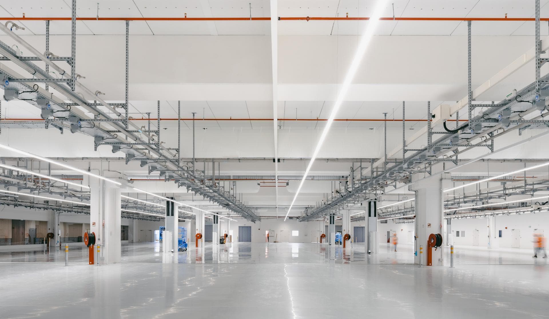 Un vaste intérieur moderne et lumineux d'un entrepôt ou d'une usine, avec un sol gris brillant et réfléchissant. Le haut plafond est traversé par des rangées de conduits métalliques complexes, de rails d'éclairage LED et de tuyaux orange, avec quelques silhouettes floues au loin.