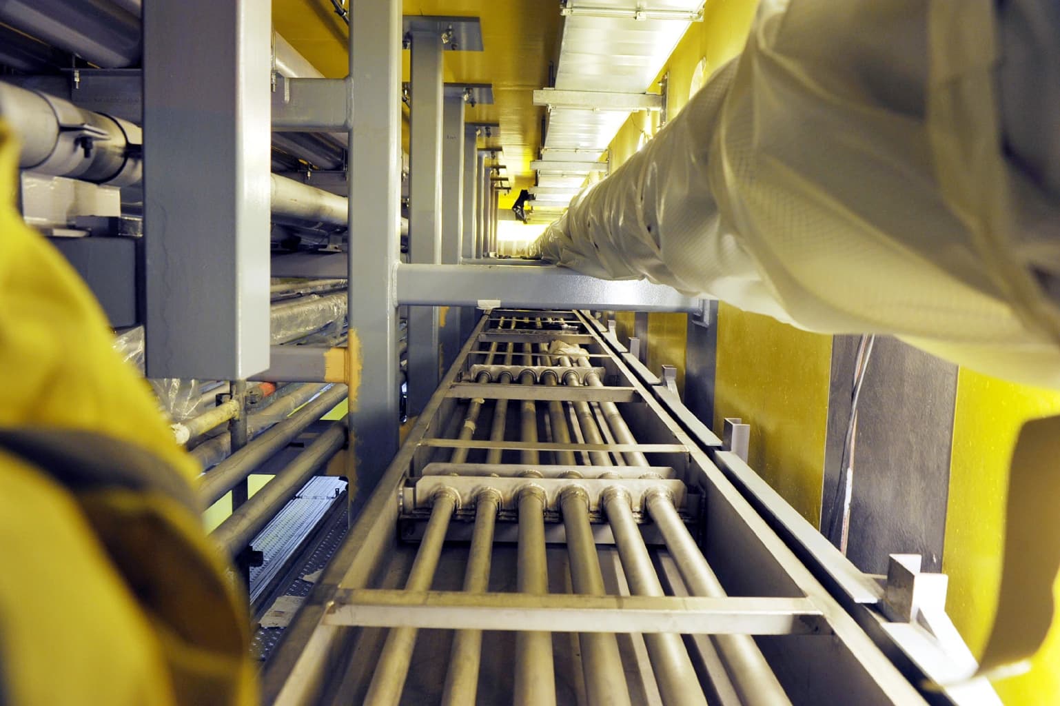 Une vue en plongée d'un canal industriel aux murs jaunes vifs, présentant de multiples tuyaux métalliques gris sur un support en rack et des poutres de soutien grises. Un grand conduit blanc en tissu s'étend le long du plafond sur la droite, créant une perspective profonde.