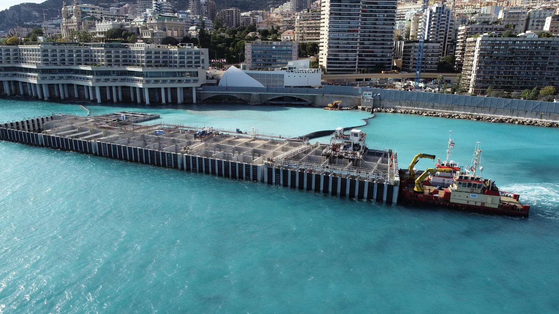 Grande structure flottante en béton, semblable à un caisson de construction avec des compartiments, déplacée par un remorqueur rouge et blanc dans des eaux turquoise. En arrière-plan, une ville côtière densément construite s'étend sur une colline.