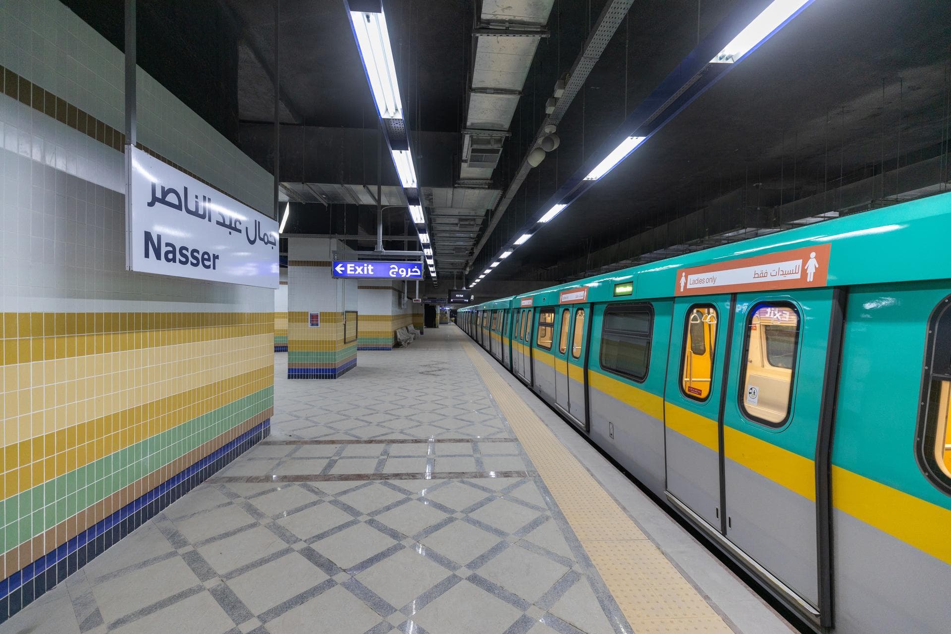 An empty, well-lit Nasser metro station platform features a teal and yellow train with a 'Ladies only' section. Tiled walls display colorful horizontal bands, complementing the grey patterned floor.