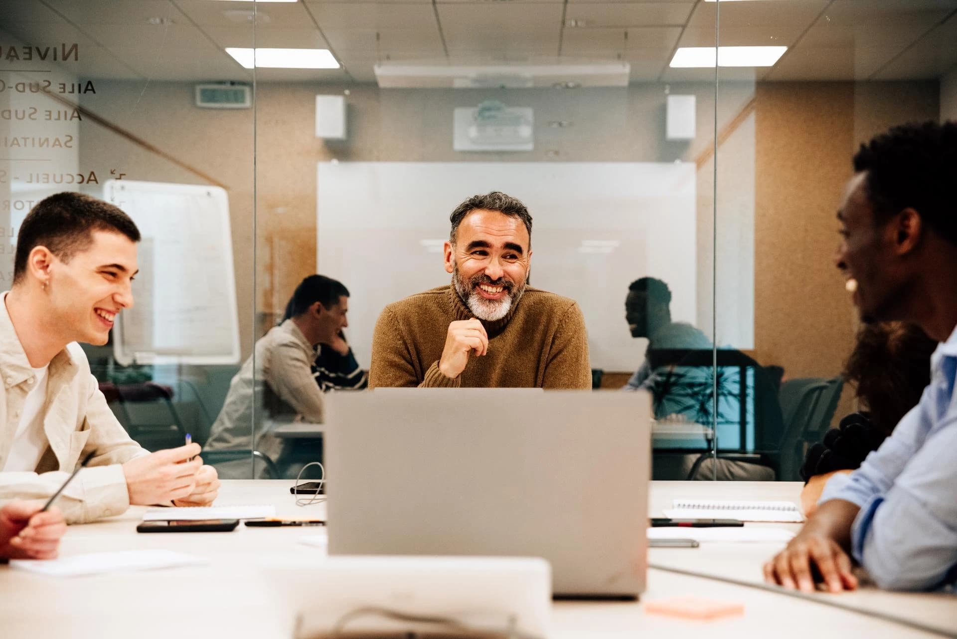 Un groupe diversifié de collègues sourit et interagit lors d'une réunion dans un bureau moderne. Un homme barbu en col roulé marron est au centre, regardant joyeusement vers la droite, avec un ordinateur portable devant lui.