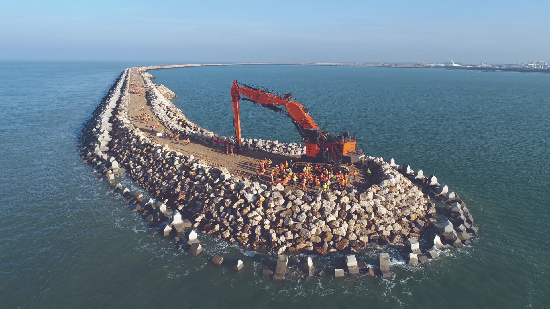 Vue aérienne d'une longue digue en construction dans la mer, où une grande pelleteuse orange et des ouvriers en gilets de sécurité jaunes et oranges travaillent sur la structure faite de rochers et de blocs de béton. La digue s'étend vers un port industriel à l'horizon, sous un ciel bleu clair.