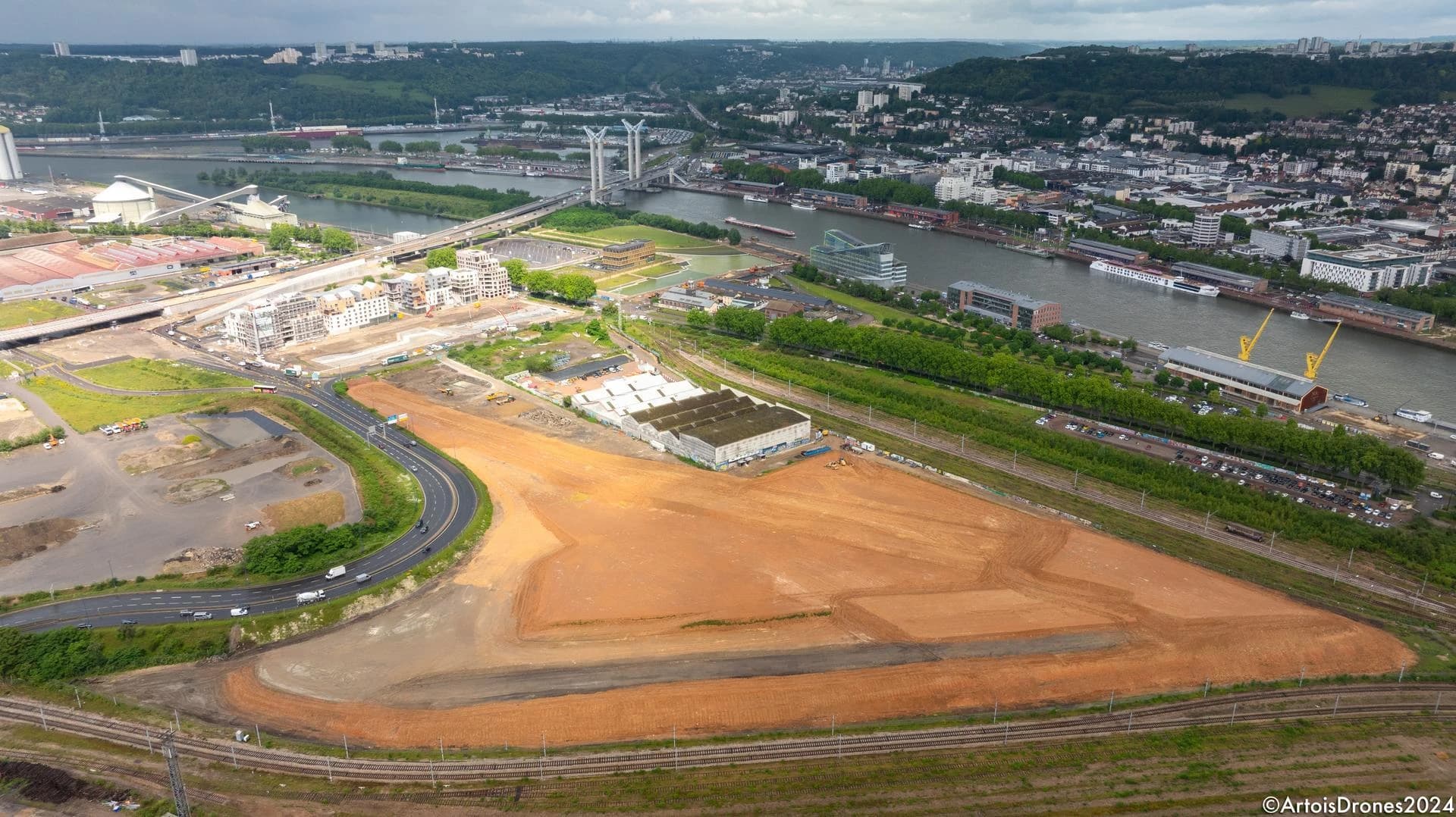 Vue aérienne d'un vaste terrain de construction orange-brun au premier plan, avec un fleuve industriel traversant une ville dense, des ponts modernes et des collines verdoyantes sous un ciel couvert.