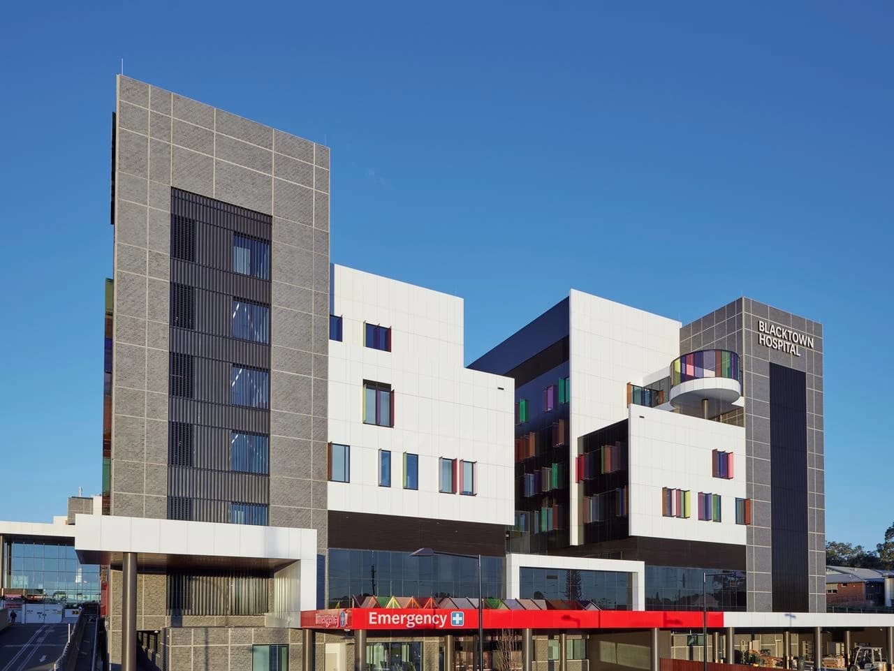 Façade extérieure moderne de l'Hôpital de Blacktown, composée de blocs gris, blancs et noirs, avec des fenêtres aux cadres colorés et un panneau rouge 'Emergency' à l'entrée.