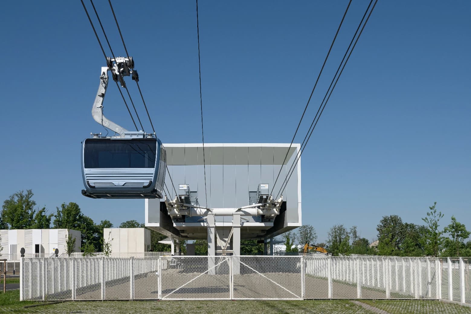 Une télécabine moderne gris et bleu avec des fenêtres teintées approche d'une station blanche et géométrique sous un ciel bleu clair. Au premier plan, une clôture en treillis blanc sépare l'herbe et le pavé, avec des arbres verts en arrière-plan.
