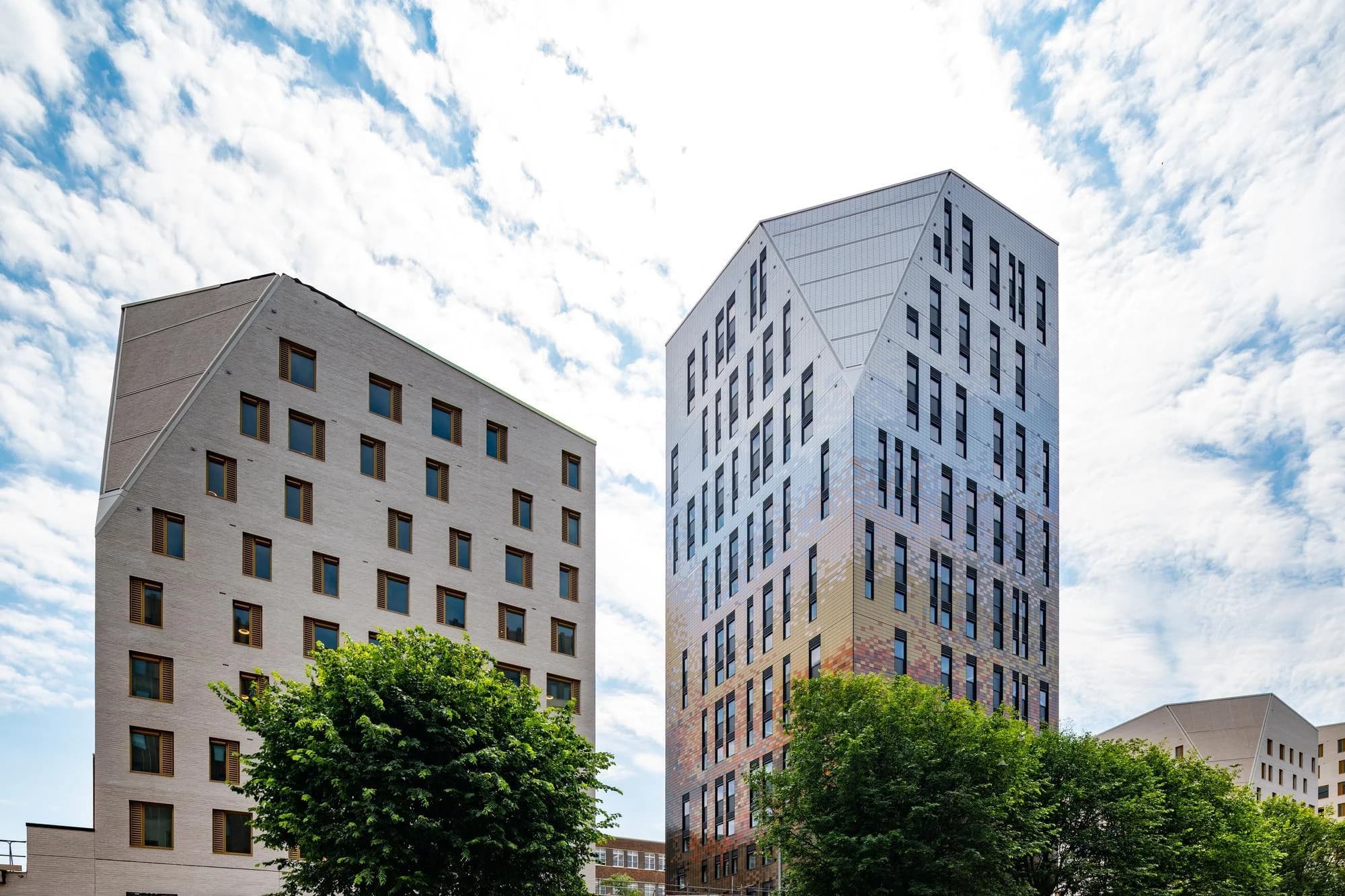 Deux bâtiments modernes se dressent sous un ciel partiellement nuageux. À gauche, une structure en briques gris clair avec des fenêtres à cadre en bois; à droite, une tour plus haute avec une façade métallique irisée passant du bleu argenté au cuivre, partiellement masquée par des arbres verts au premier plan.