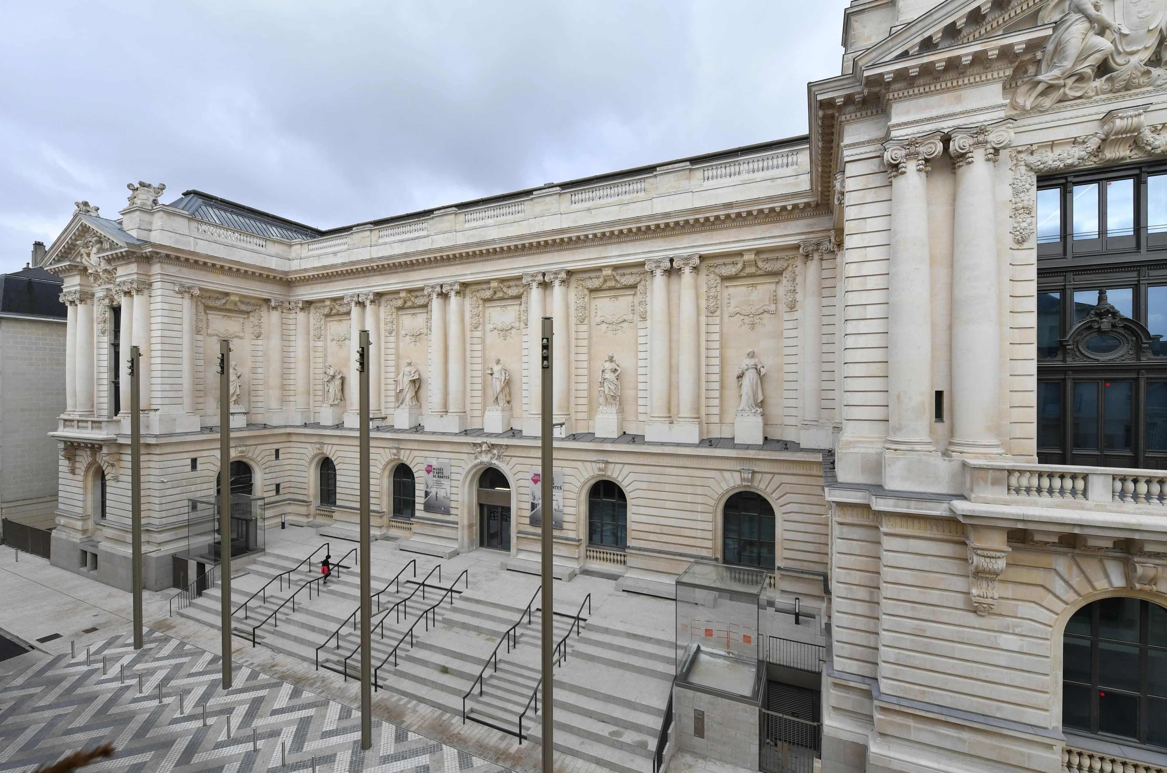 Façade imposante et ornée du Musée des Arts de Nantes en pierre claire sous un ciel nuageux, avec des colonnes, statues et fenêtres arquées. Un large escalier en béton avec des rampes noires mène aux entrées, donnant sur un parvis moderne au motif géométrique gris.