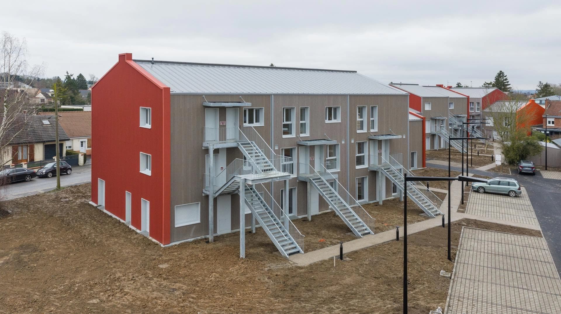 Bâtiment moderne avec un mur rouge, bardage bois et escaliers extérieurs en métal en zone résidentielle.