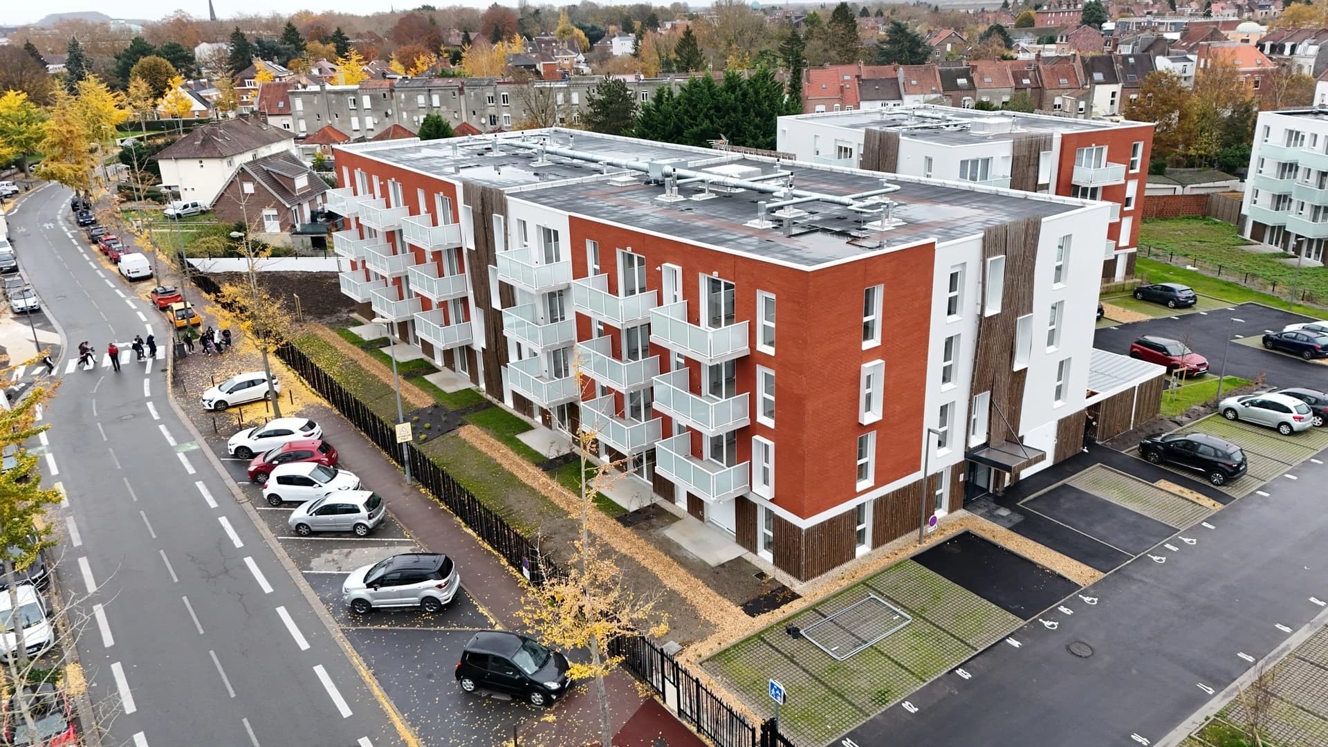 Immeuble moderne avec briques rouges, balcons blancs et parking, situé dans un environnement urbain.