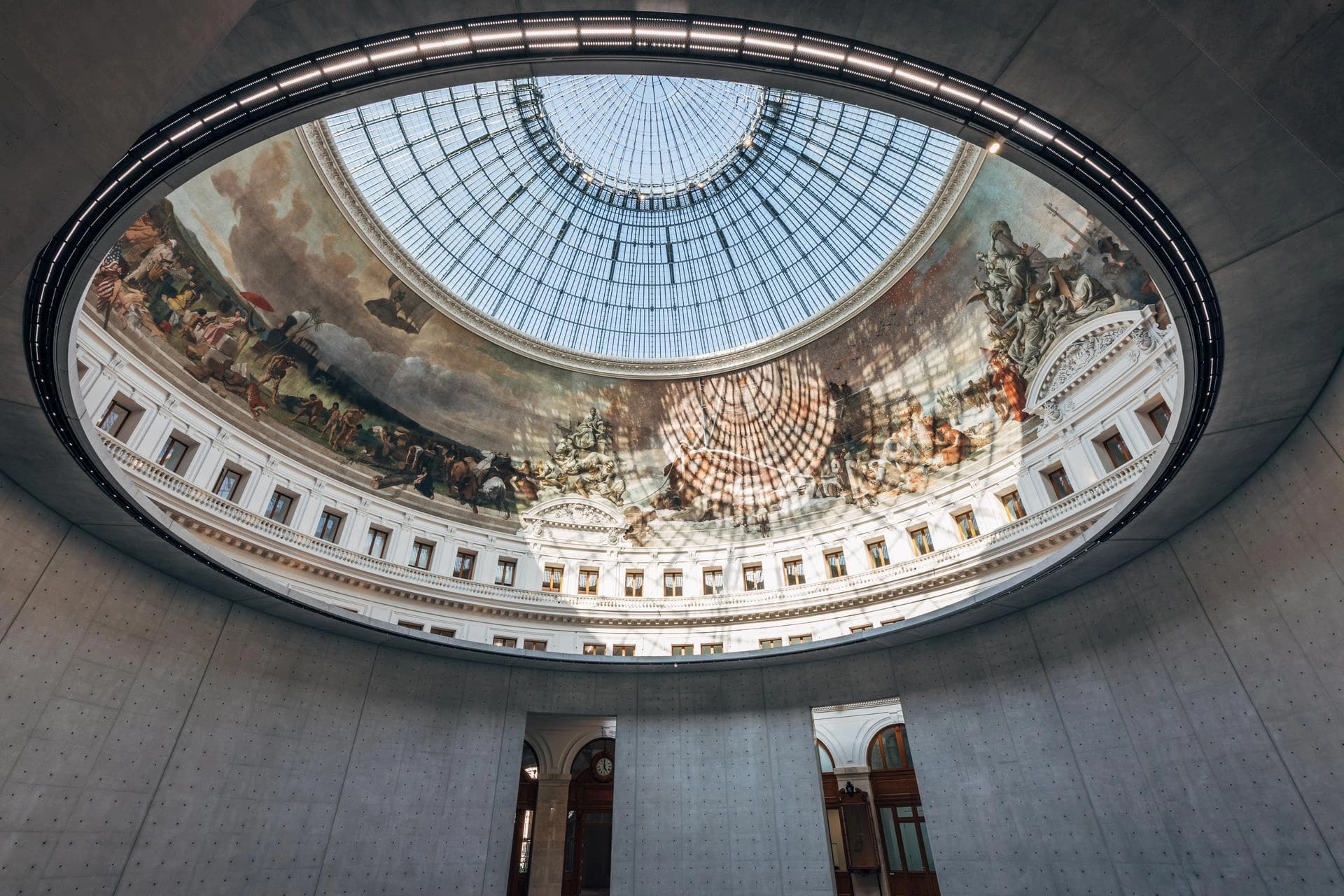 Vue de l'intérieur de la Bourse de Commerce montrant une coupole vitrée entourée d'une grande fresque circulaire aux couleurs vives, baignée de lumière naturelle projetant des ombres graphiques. Des murs modernes en béton gris contrastent avec l'architecture classique visible sous la fresque.