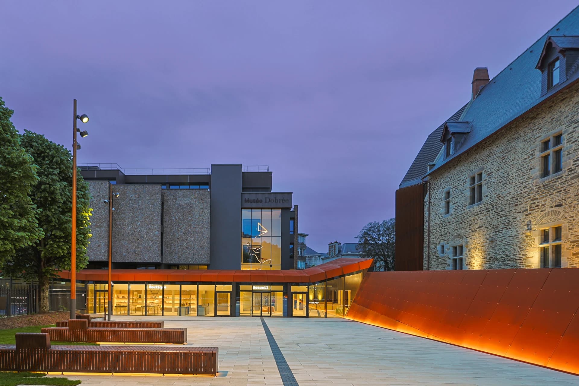 Vue extérieure du Musée Dobrée au crépuscule, combinant un bâtiment historique en pierre et une extension moderne gris et verre, avec un parvis lumineux doté de bancs et de murs orange-rouille sous un ciel teinté de violet.