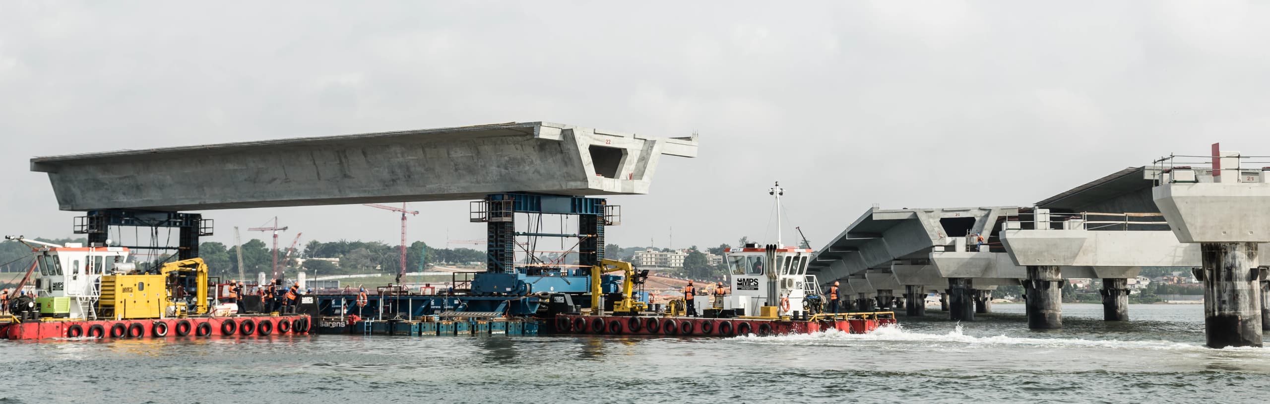 Des segments de pont en béton gris sont installés sur l'eau, avec des barges de construction, dont une avec des machines jaunes et des ouvriers en gilets orange, déplaçant un segment vers une partie du pont déjà érigée sous un ciel couvert.