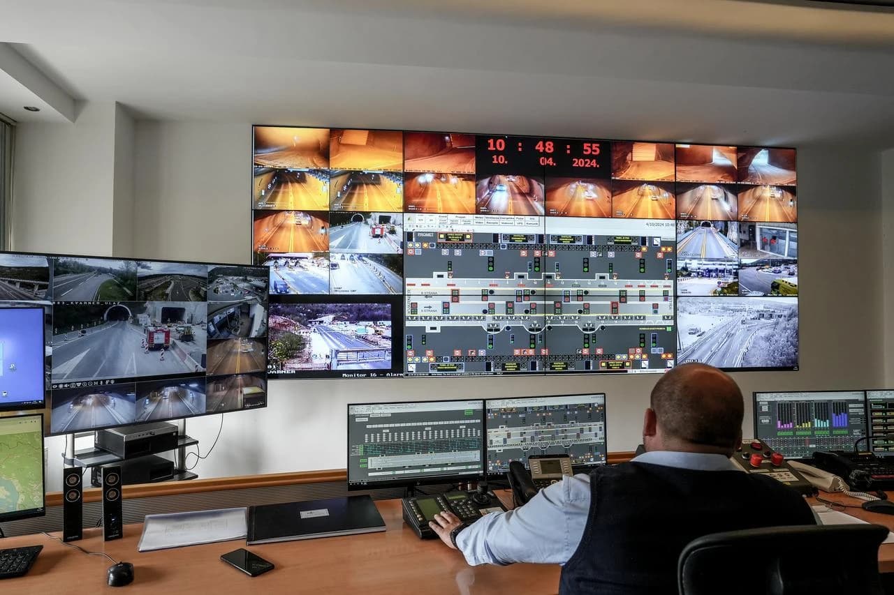 Un homme chauve est assis à un bureau dans une salle de contrôle, faisant face à un grand mur vidéo affichant de multiples flux de caméras en direct de routes, de tunnels et de chantiers de construction, ainsi que des diagrammes de systèmes numériques. Son bureau est équipé d'écrans supplémentaires, de panneaux de contrôle et de dispositifs de communication.