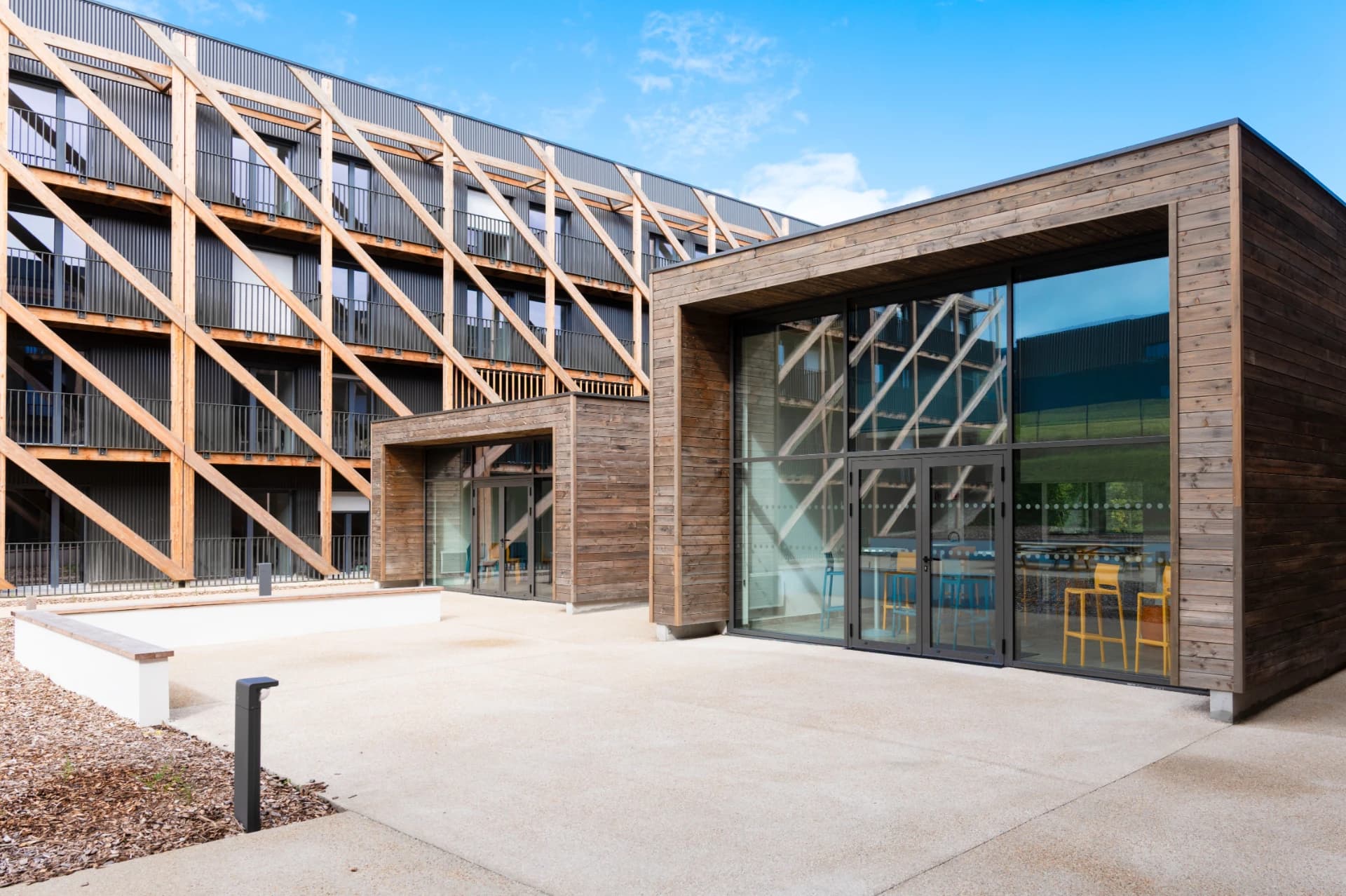 Bâtiment moderne présentant une façade multi-étages avec une structure diagonale en bois clair sur un bardage métallique sombre, juxtaposé à une section inférieure en bois foncé dotée de grandes baies vitrées, le tout donnant sur une cour en béton clair sous un ciel bleu.