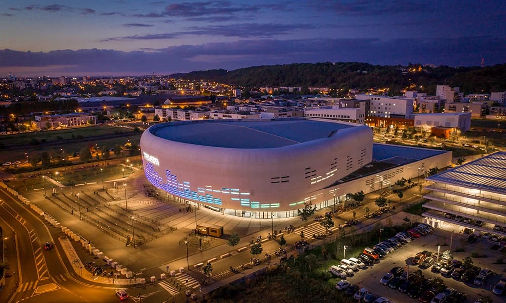 Vue aérienne de la moderne K PARC Arena aux courbes blanches, sa façade illuminée de lumières bleues et vertes vives, au crépuscule. L'arène est entourée d'un paysage urbain éclairé par les lumières de la ville et dominé par une colline boisée à l'arrière-plan.