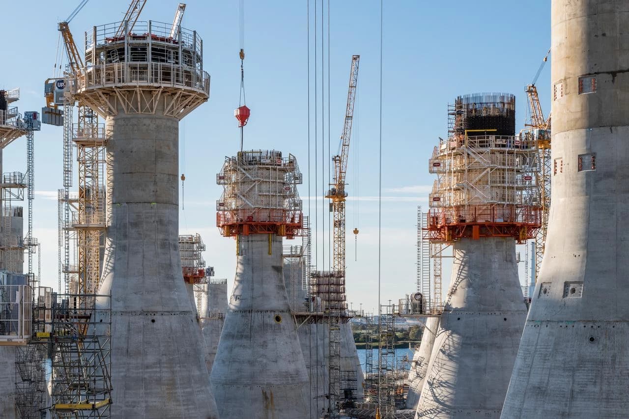 Un chantier de construction montre de grands piliers de béton gris inachevés, surmontés d'échafaudages et de coffrages, sous un ciel bleu clair avec plusieurs grues jaunes actives et un plan d'eau en arrière-plan.