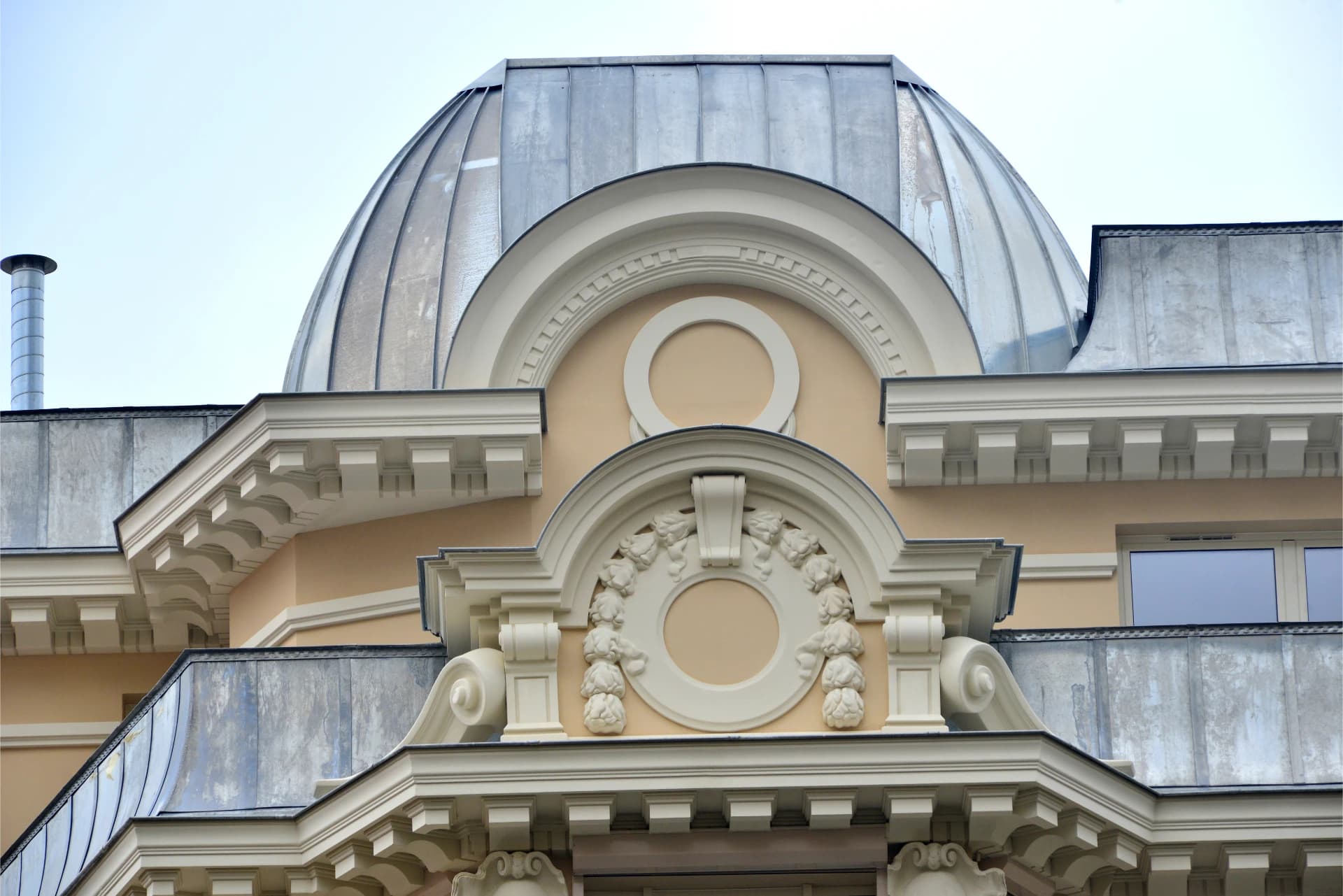 Détail architectural d'un bâtiment beige orné, présentant un grand dôme métallique argenté, des frontons cintrés blancs et des motifs sculptés floraux autour de médaillons, sous un ciel pâle.