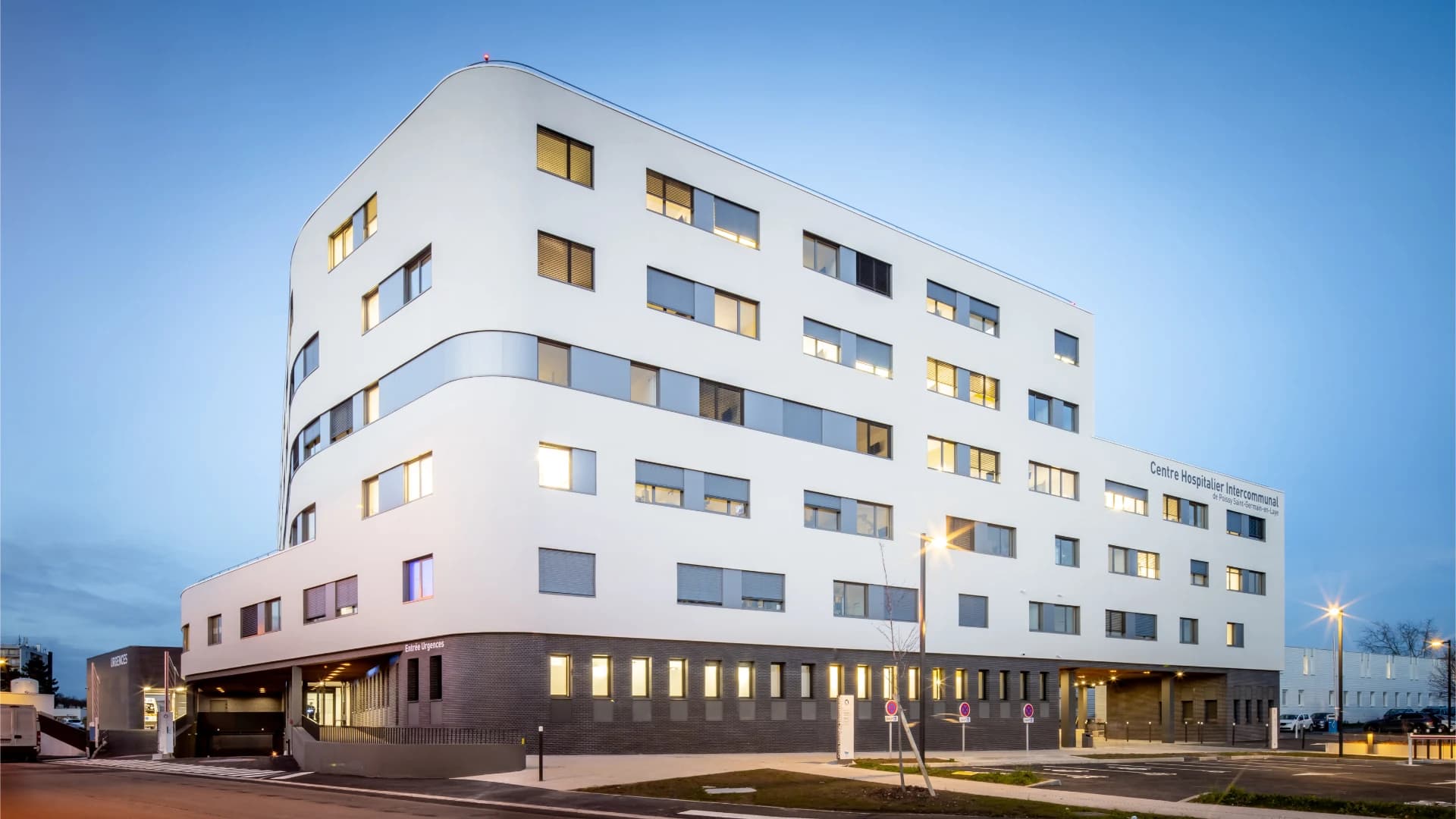 Vue extérieure d'un Centre Hospitalier Intercommunal moderne et blanc avec des façades incurvées, de nombreuses fenêtres éclairées et une entrée d'urgences visible, sous un ciel de crépuscule.