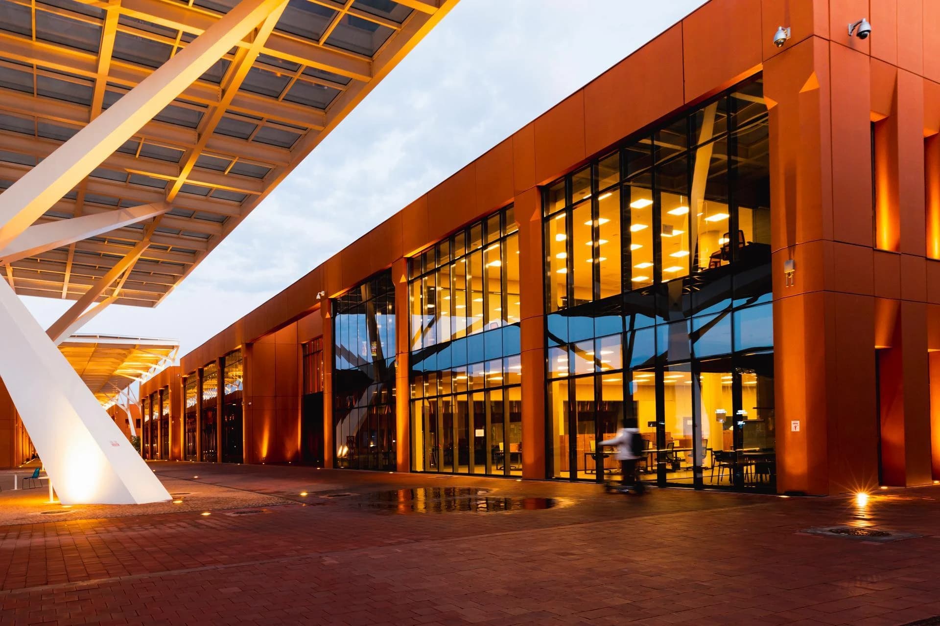 Bâtiment moderne avec une façade orange vif et de grandes baies vitrées reflétant le ciel du crépuscule, éclairées par une lumière intérieure chaleureuse. Une large structure architecturale blanche et anguleuse couvre un trottoir en briques rouges mouillées, où une silhouette floue passe.