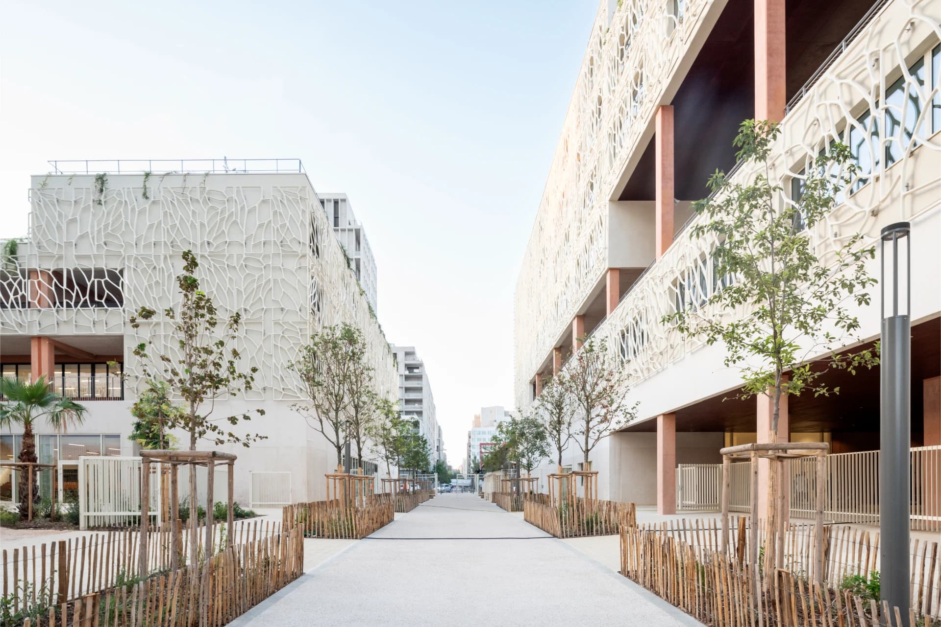 Un chemin piétonnier moderne est flanqué de deux bâtiments aux façades claires ornées de motifs organiques en treillis, avec des balcons couleur terre cuite et des rangées de jeunes arbres bordés de clôtures en bois. Le chemin clair mène vers l'horizon sous un ciel lumineux.