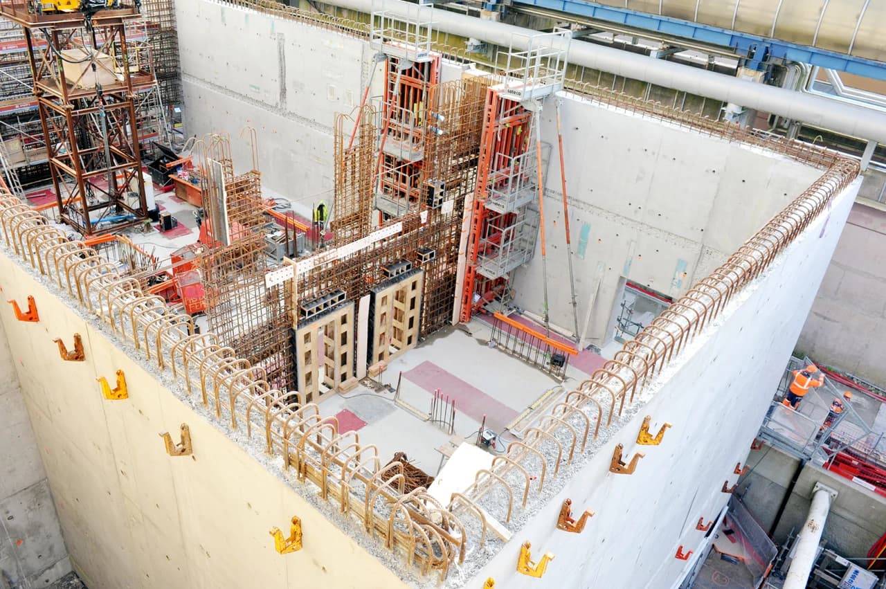 Vue plongeante sur un vaste chantier de construction, présentant des murs massifs en béton gris avec des armatures en acier courbé, des échafaudages complexes orange et gris, et des ouvriers s'affairant à la construction de structures internes.