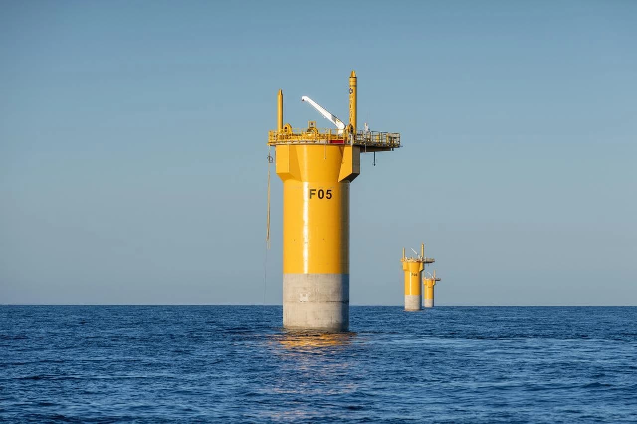 Un grand flotteur offshore jaune vif, marqué "F05" sur son corps cylindrique et doté d'une base en béton gris et d'une plateforme d'équipement, se dresse dans la mer bleue calme sous un ciel clair. Plusieurs structures jaunes similaires plus petites sont visibles plus loin dans l'eau sereine.