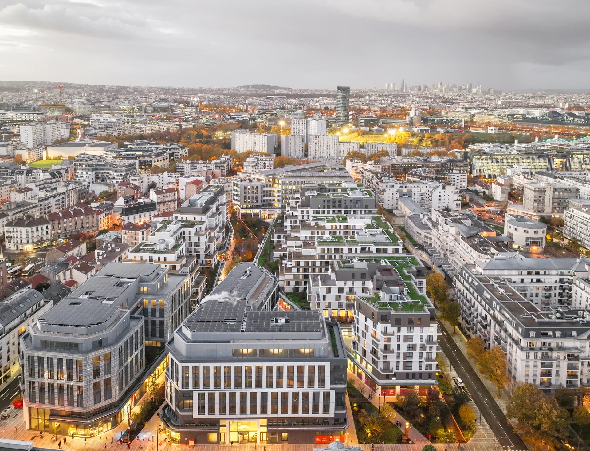 Vue aérienne d'un quartier urbain moderne au crépuscule, présentant de nombreux immeubles récents avec toits végétalisés et panneaux solaires, leurs fenêtres et les rues étant éclairées de teintes chaudes sous un ciel gris nuageux, avec l'horizon lointain de la ville.