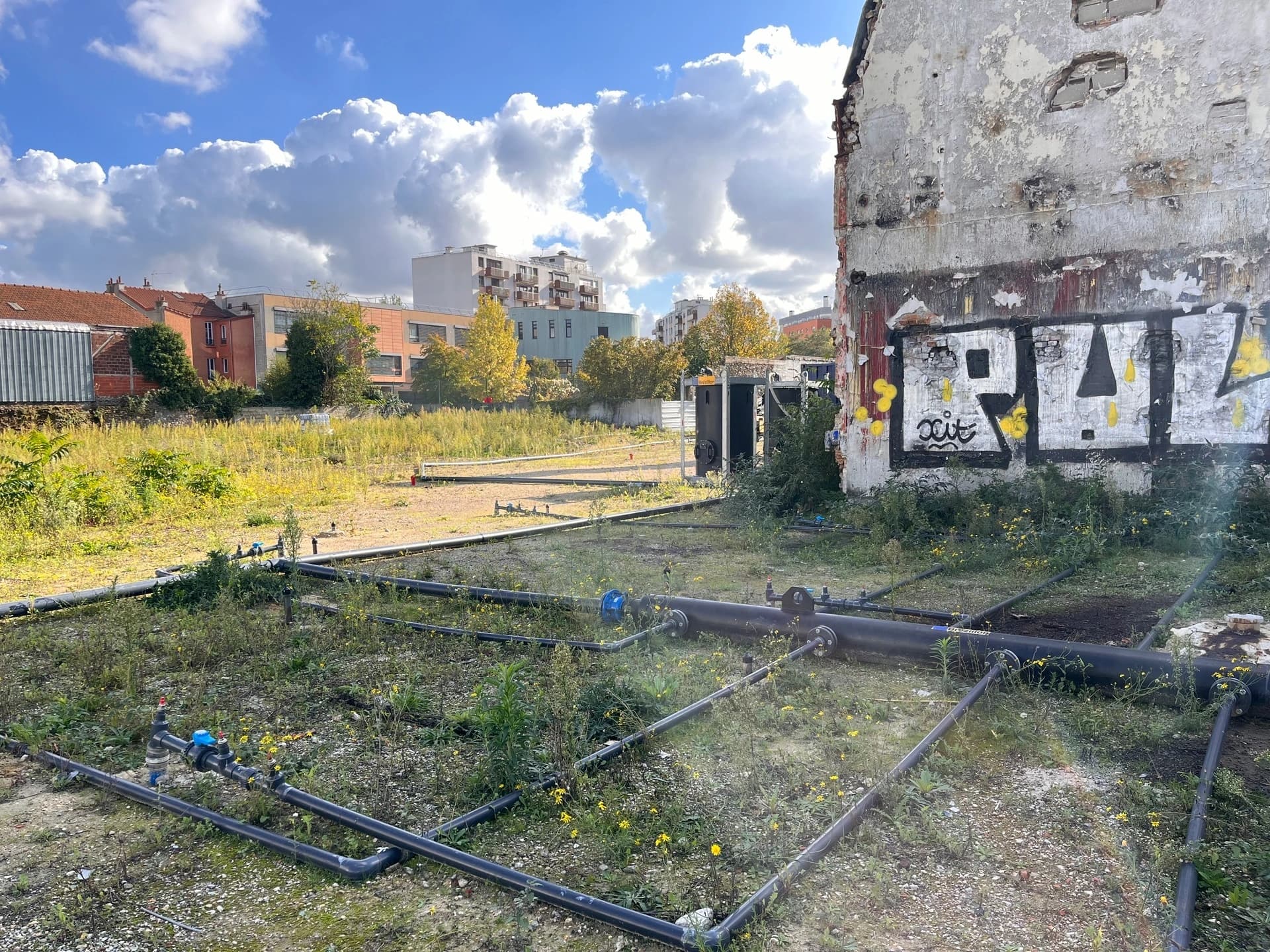 Vue d'un terrain vague urbain avec un réseau de tuyaux noirs et des valves bleues au premier plan, devant un bâtiment délabré et graffé, et des immeubles résidentiels modernes en arrière-plan sous un ciel bleu nuageux.