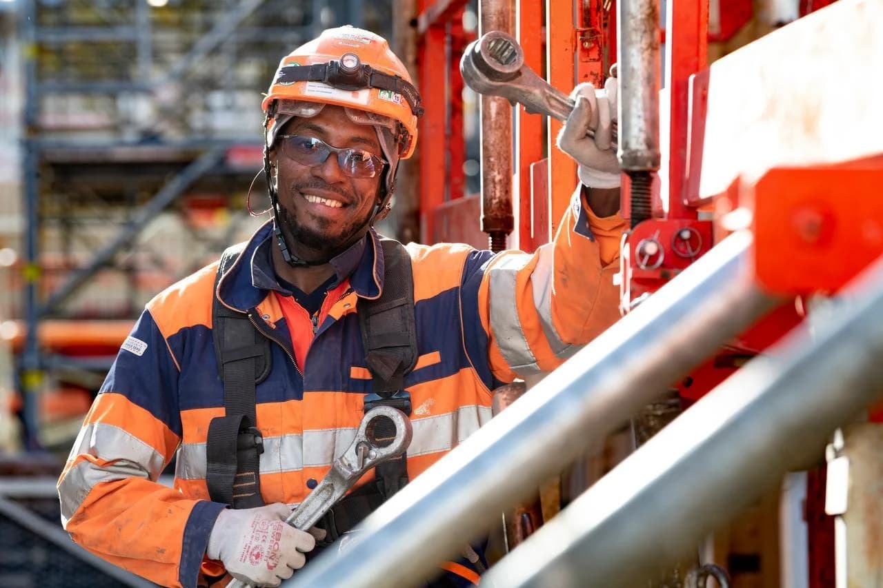 Un homme noir souriant, coiffé d'un casque orange et de lunettes de sécurité, porte une veste de travail haute-visibilité orange et bleue, tenant des clés à molette sur un chantier de construction.