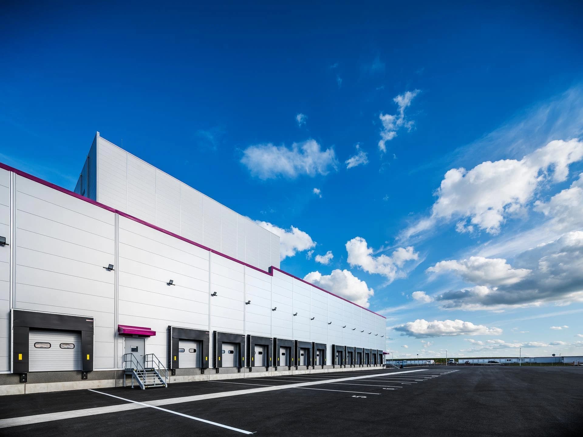 Un grand entrepôt logistique moderne aux façades blanches et argentées présente une longue rangée de quais de chargement noirs et des touches de violet. Le tout est sous un ciel bleu lumineux avec des nuages blancs, face à un vaste parking asphalté vide.