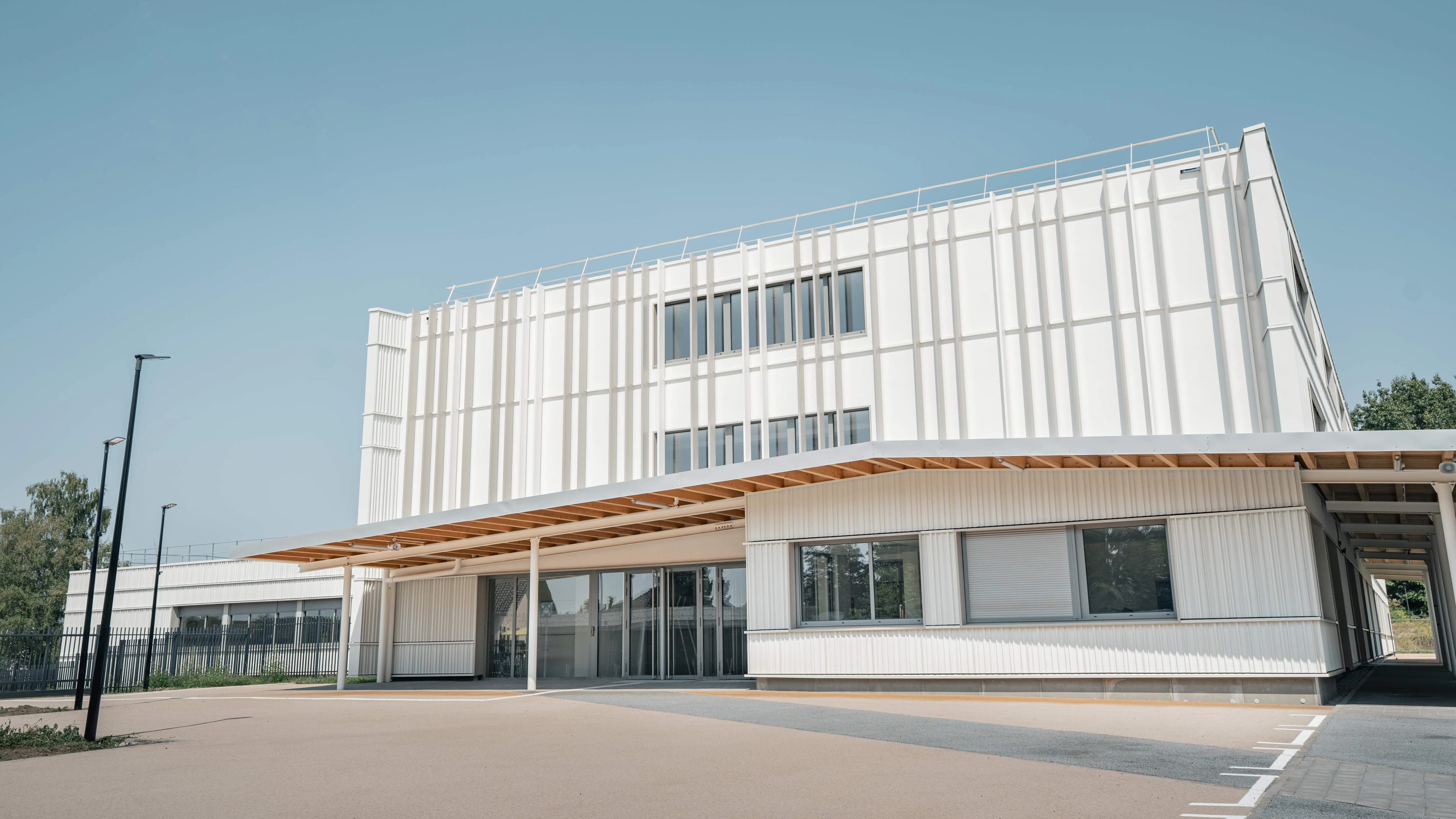 Un bâtiment moderne blanc à deux étages se dresse sous un ciel bleu clair, avec une façade distincte de panneaux verticaux et de grandes fenêtres. Une large marquise avec des poutres en bois apparentes s'étend au-dessus de l'entrée du rez-de-chaussée, devant un parvis pavé beige et gris.
